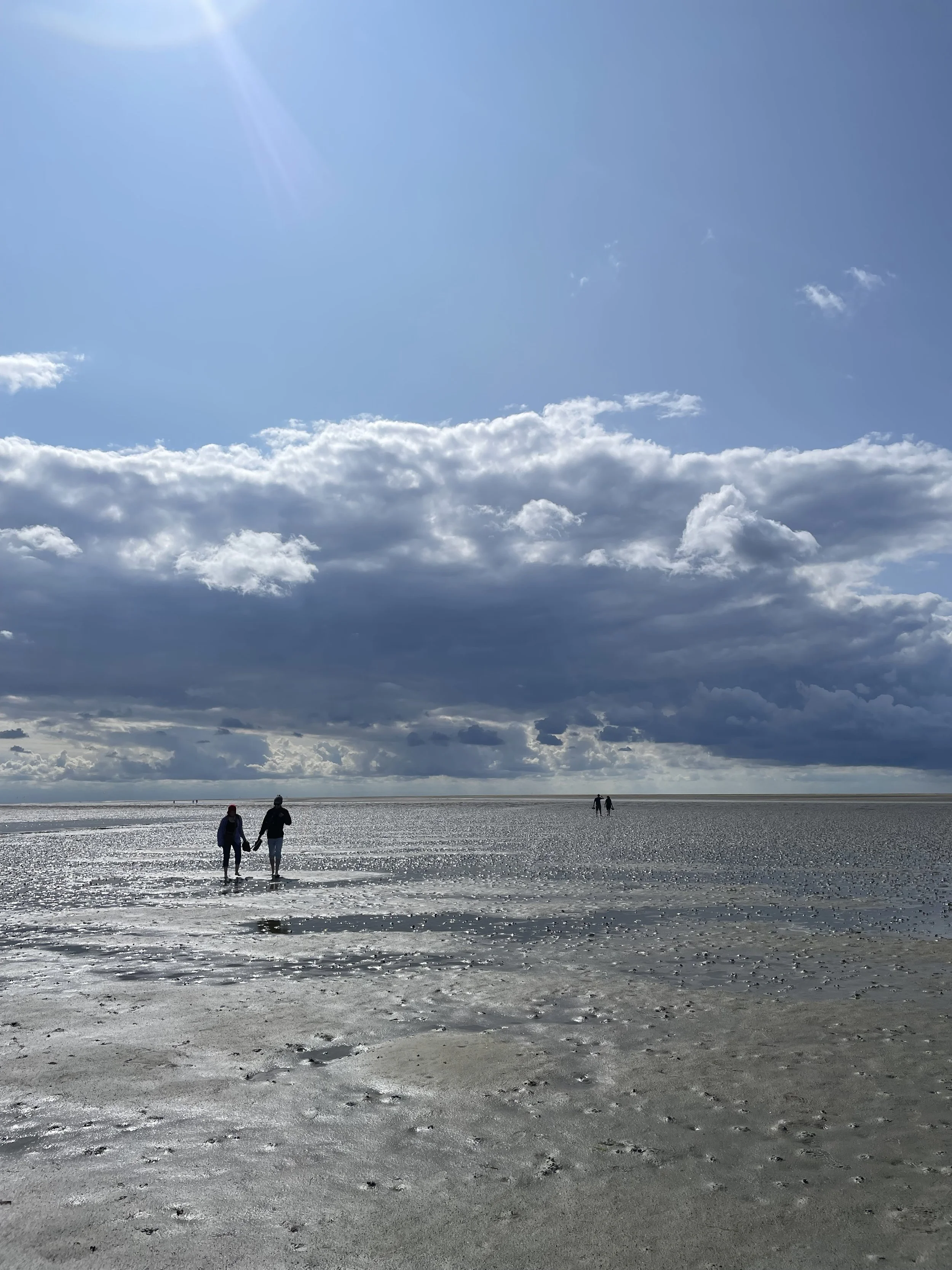 Menschen, die am Strand entlang spazieren, mit einem dramatischen Himmel voller Wolken und Sonnenstrahlen, spiegelndes Wasser auf dem Sand.