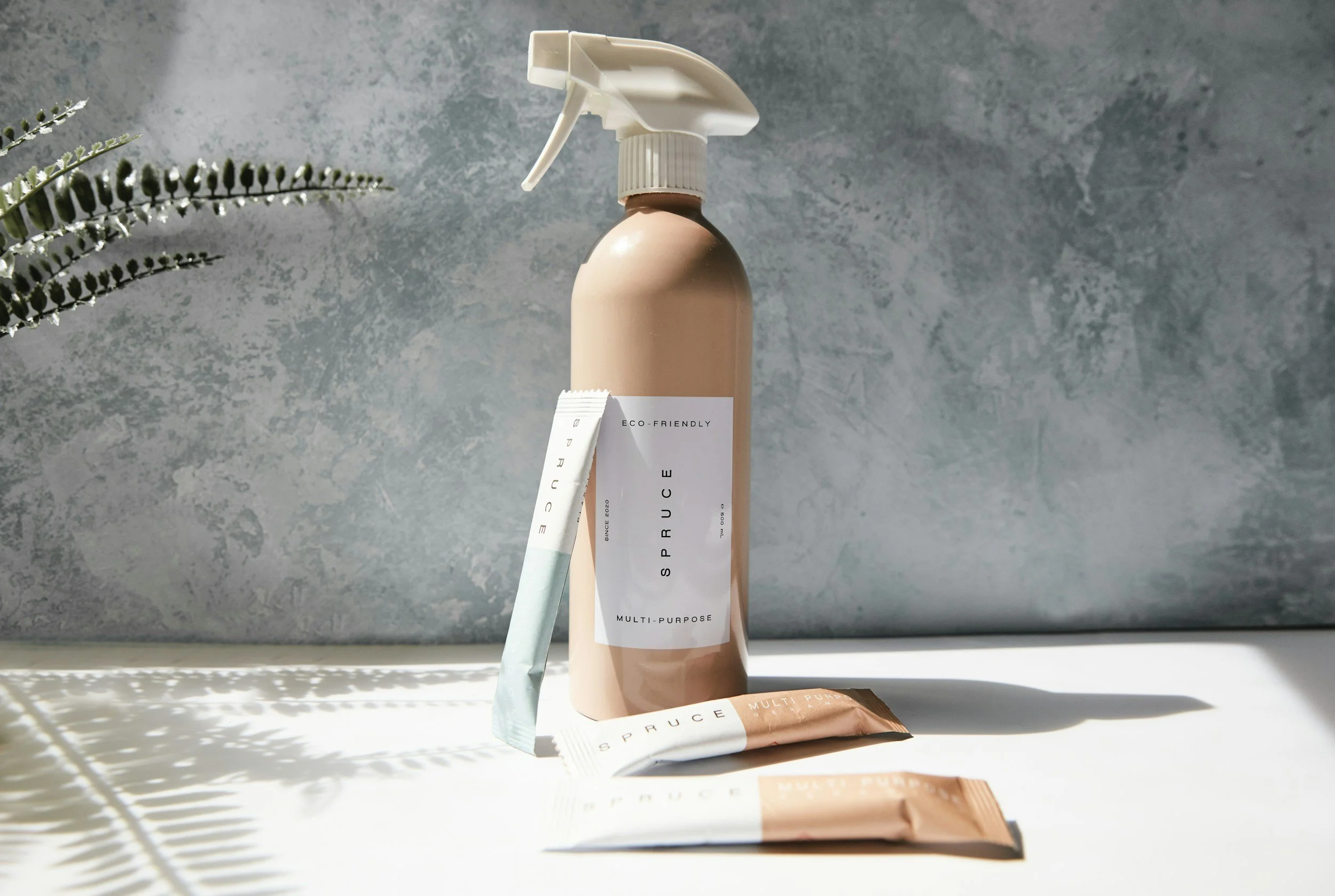 A beige spray bottle labeled 'Spruce Multi-Purpose Eco-Friendly' with two sachets on a white surface, with sunlight casting shadows and a green plant left of the image against a textured blue wall.