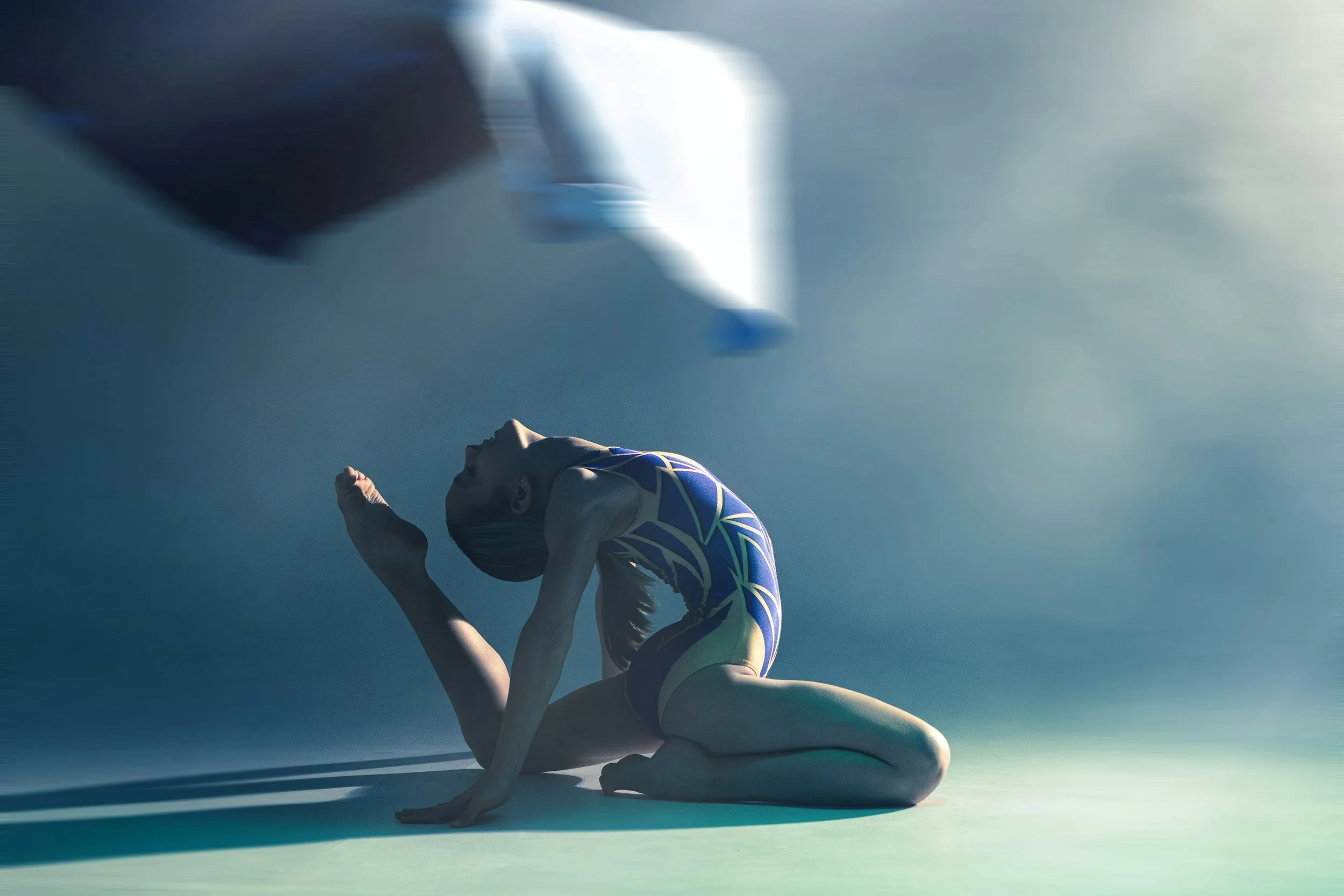A gymnast in a colorful leotard performs a yoga pose on a light-colored floor with a gradient blue background.