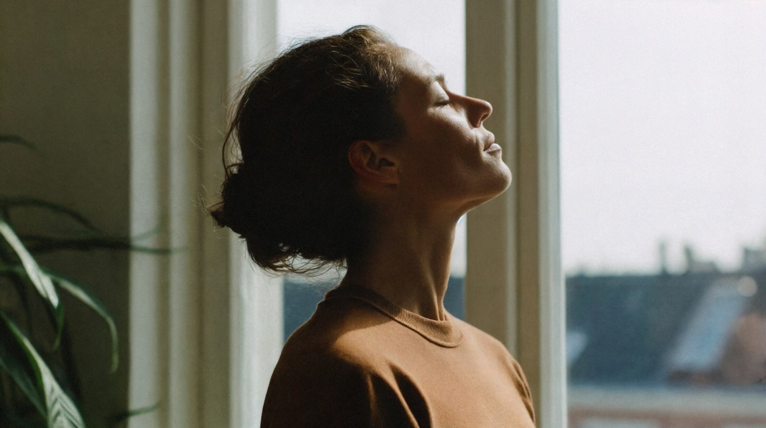 A woman with short hair laying her head back, eyes closed, facing a window with soft natural light.
