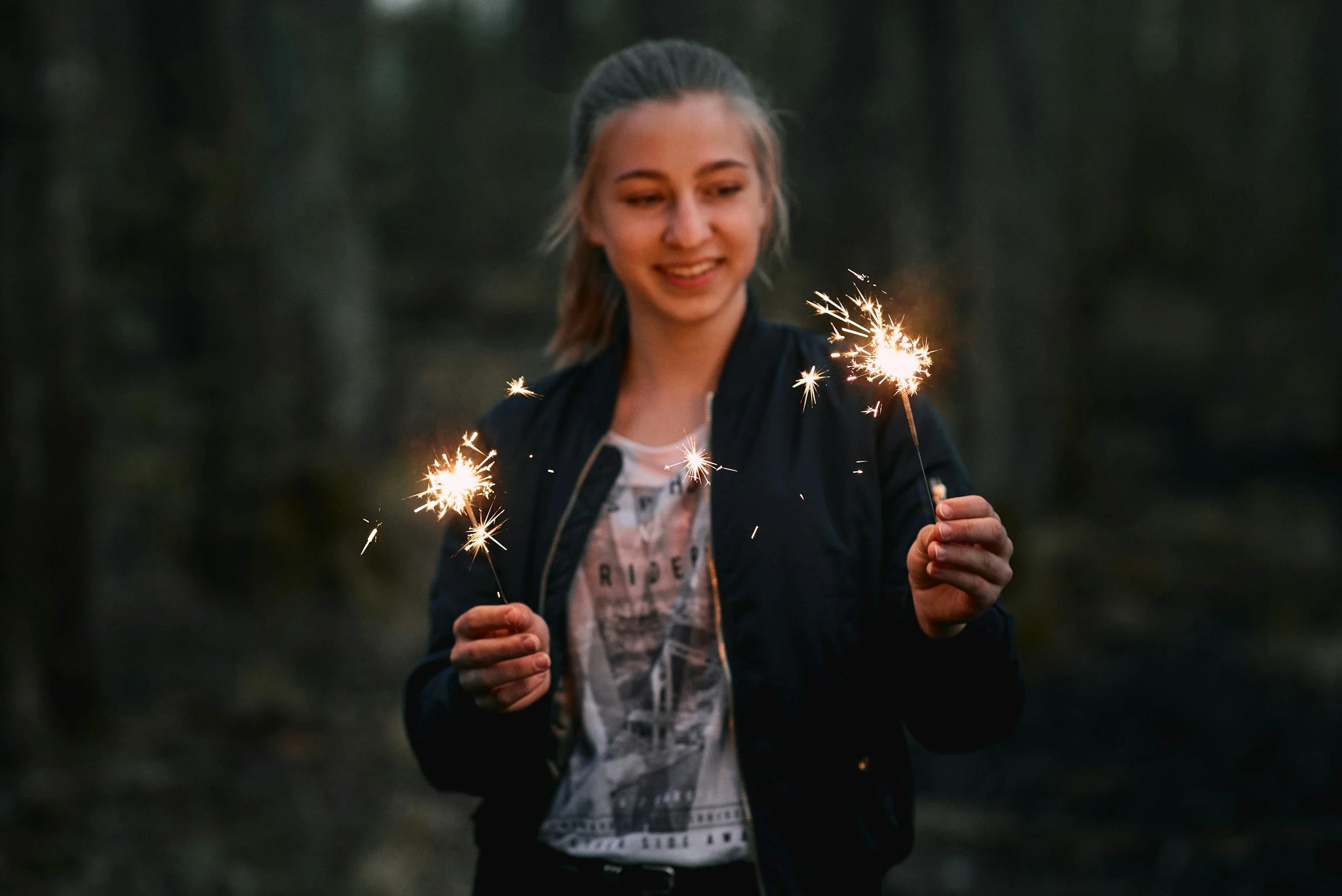 A young woman outdoors holding sparklers and smiling, with a dark forest background.
