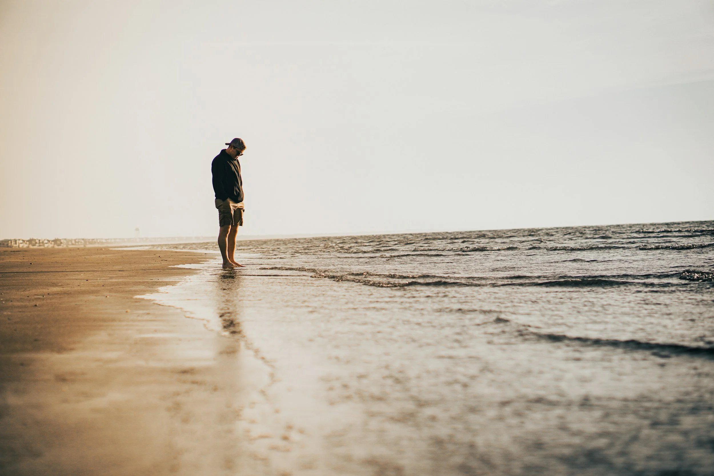 A person standing at the edge of the ocean, wearing a hat, black jacket, and shorts, looking down at the water in a peaceful, natural setting during sunset or sunrise.