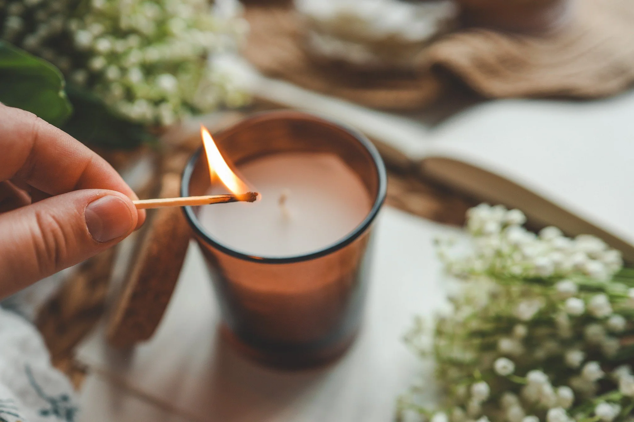 Hand holding a lit matchstick near a candle in a brown glass holder with flowers and decorative objects in the background.