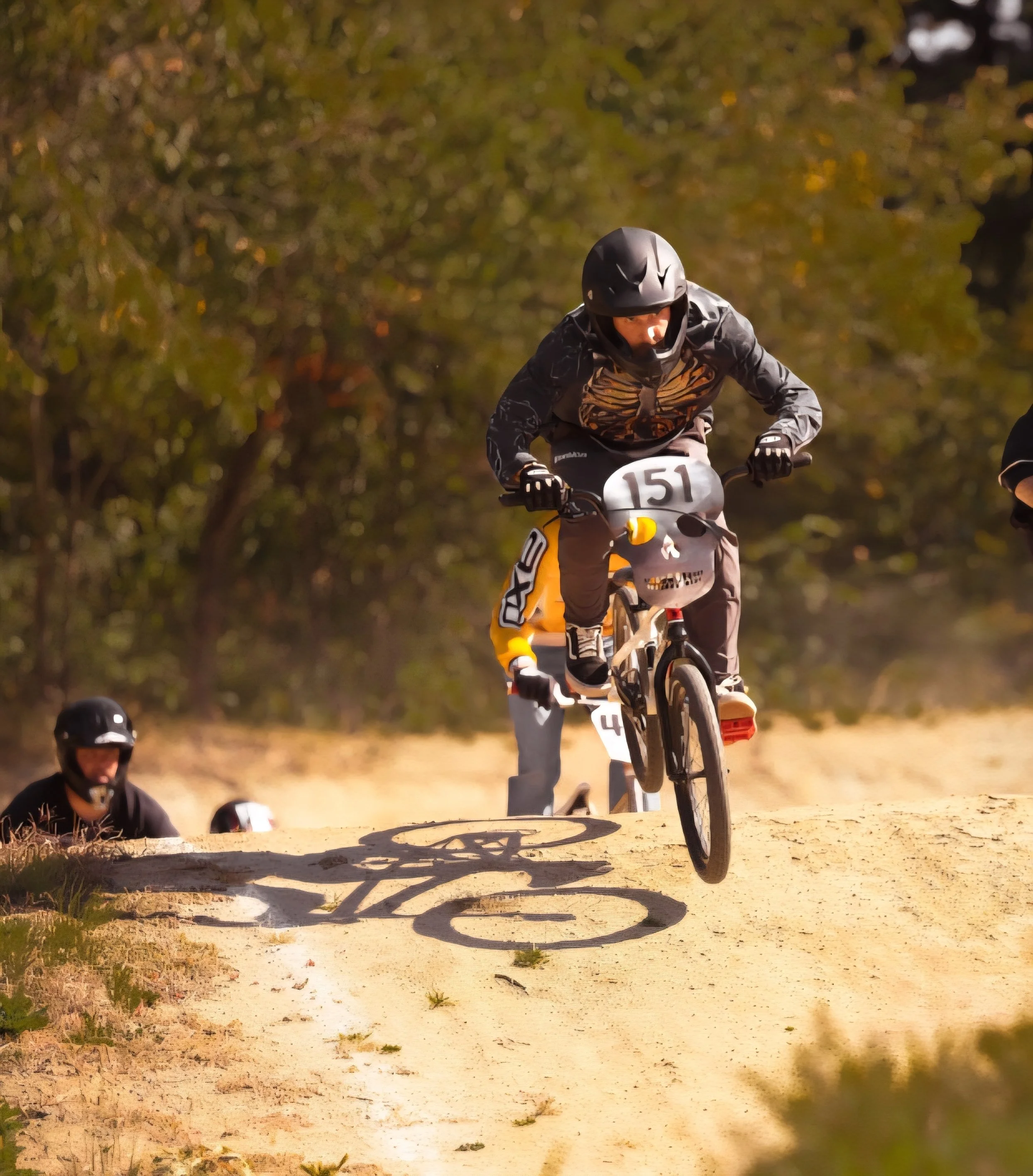 A mountain biker in mid-air jumping over a dirt trail, wearing a helmet and sports gear, with trees and other riders in the background.