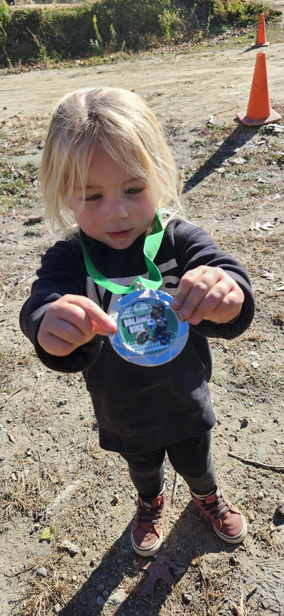 A young girl with blonde hair wearing a black hoodie, black leggings, and red sneakers, holding up a medal with a green ribbon, outdoors on a dirt area with orange traffic cones in the background.
