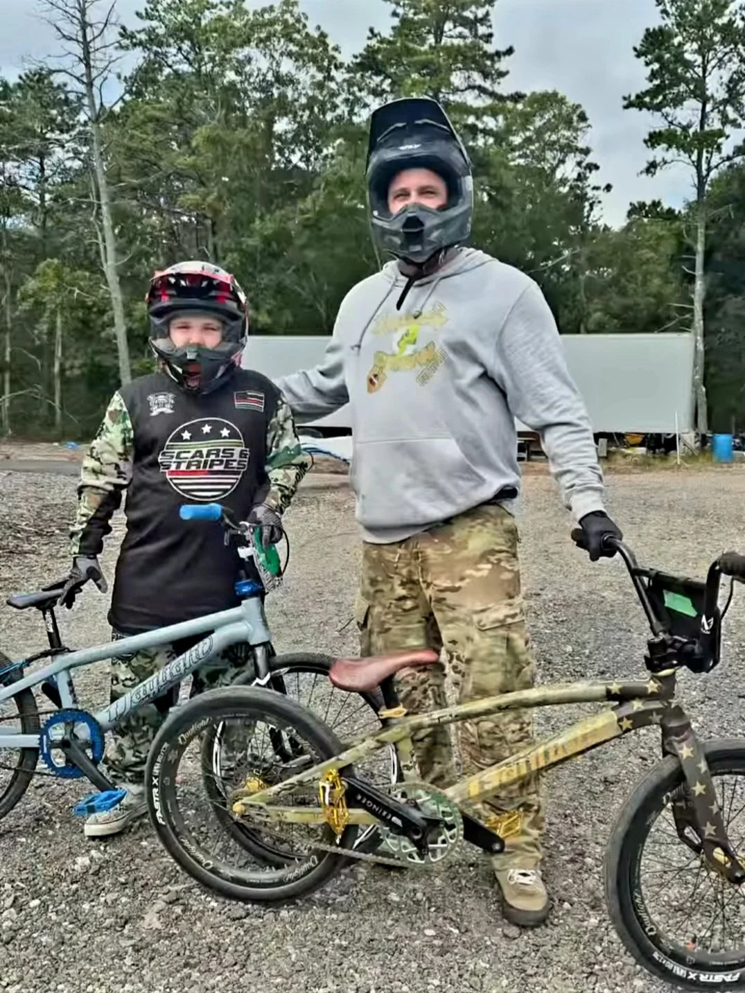 A man and a young boy standing outdoors with their bikes, both wearing helmets and gear, smiling at the camera.