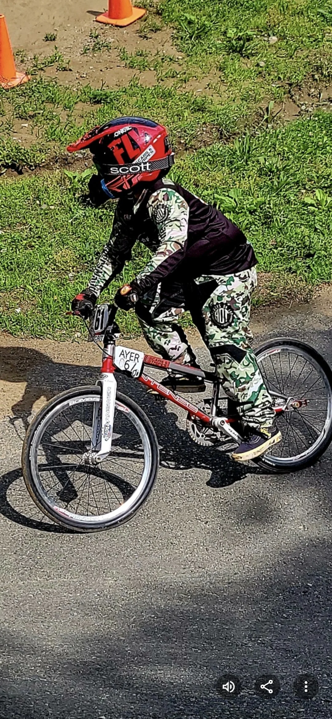 A young person riding a mountain bike on a paved road with grass and orange traffic cones in the background. The rider is wearing a red helmet with black and white details, a black long-sleeve shirt, and camouflage pants.