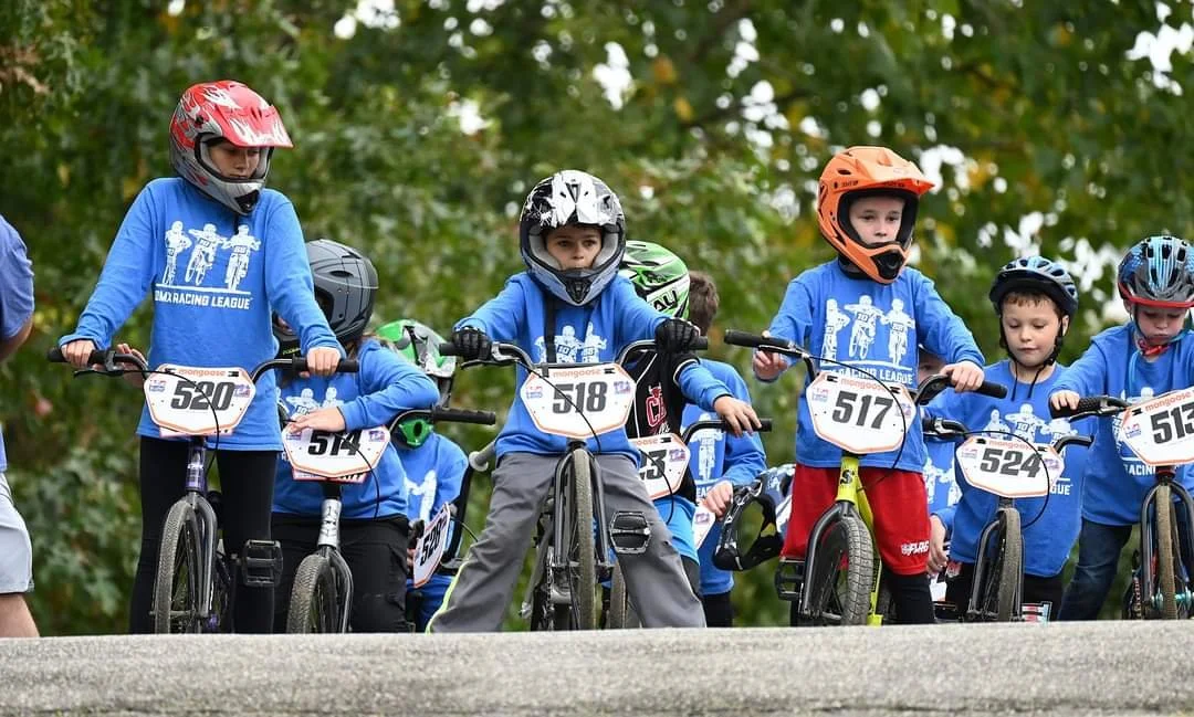 Children in blue shirts with race numbers on bikes, wearing helmets, preparing for a bike race outdoors