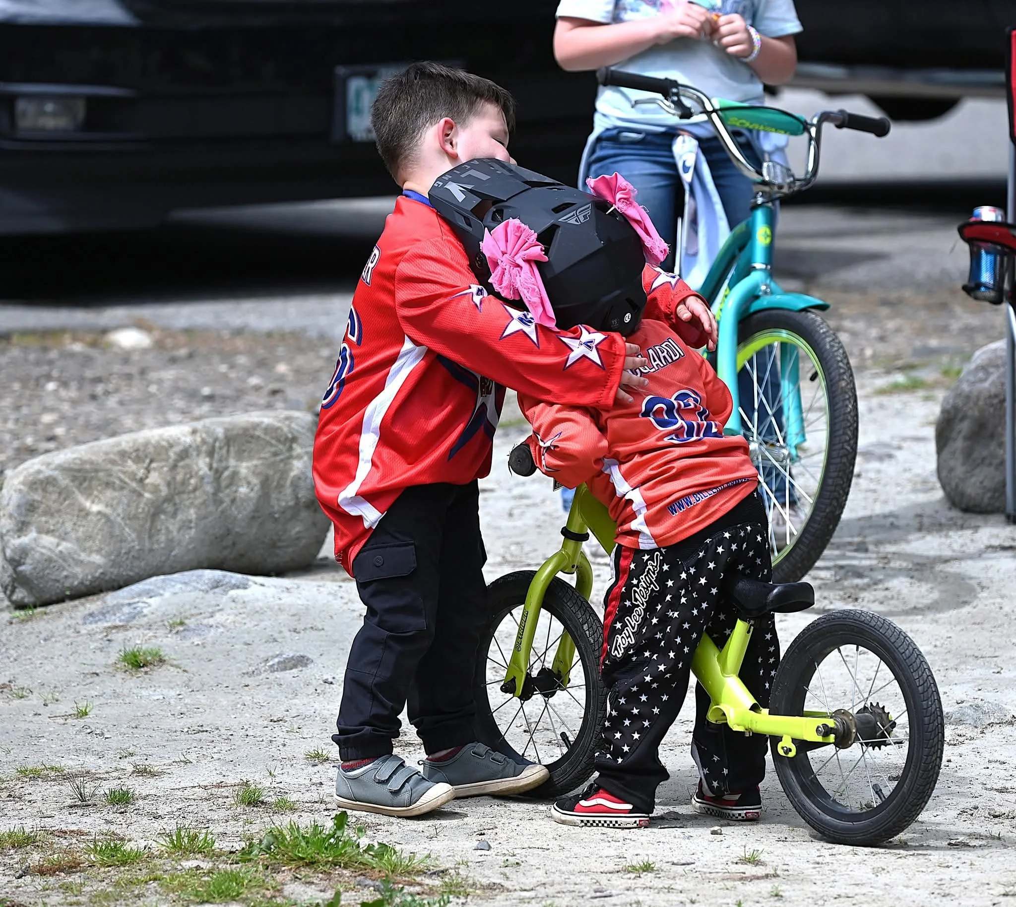 Two boys, one with a helmet, are interacting with a small green balance bike outdoors near rocks. One boy in a red hockey jersey helps the other who is sitting on the bike. A woman with a teal bicycle and a jacket tied to her waist is visible in the background.