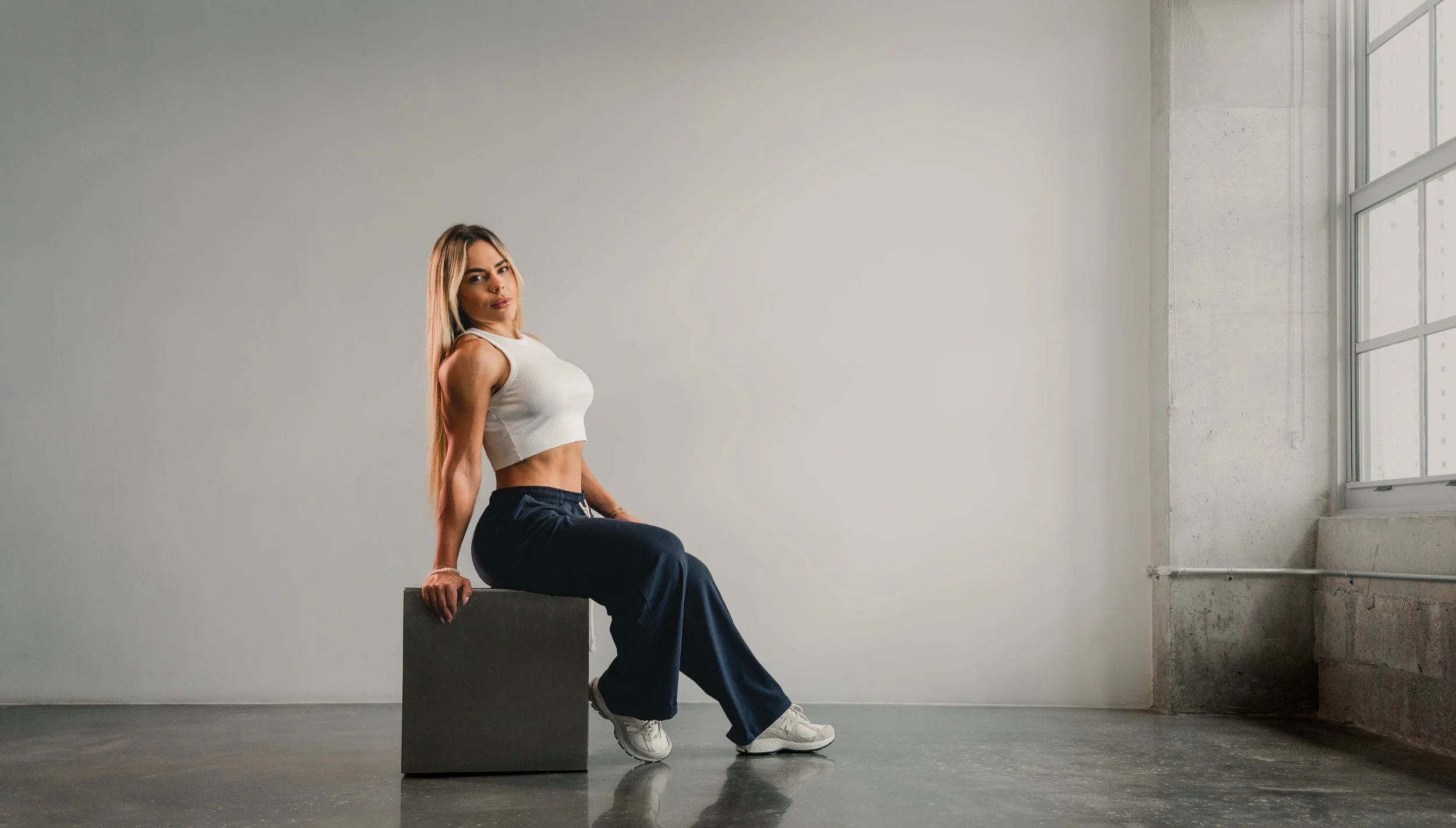 A woman with long blonde hair sitting on a gray block in an empty room with large windows and concrete walls, wearing a white sleeveless crop top, dark blue pants, and white sneakers.