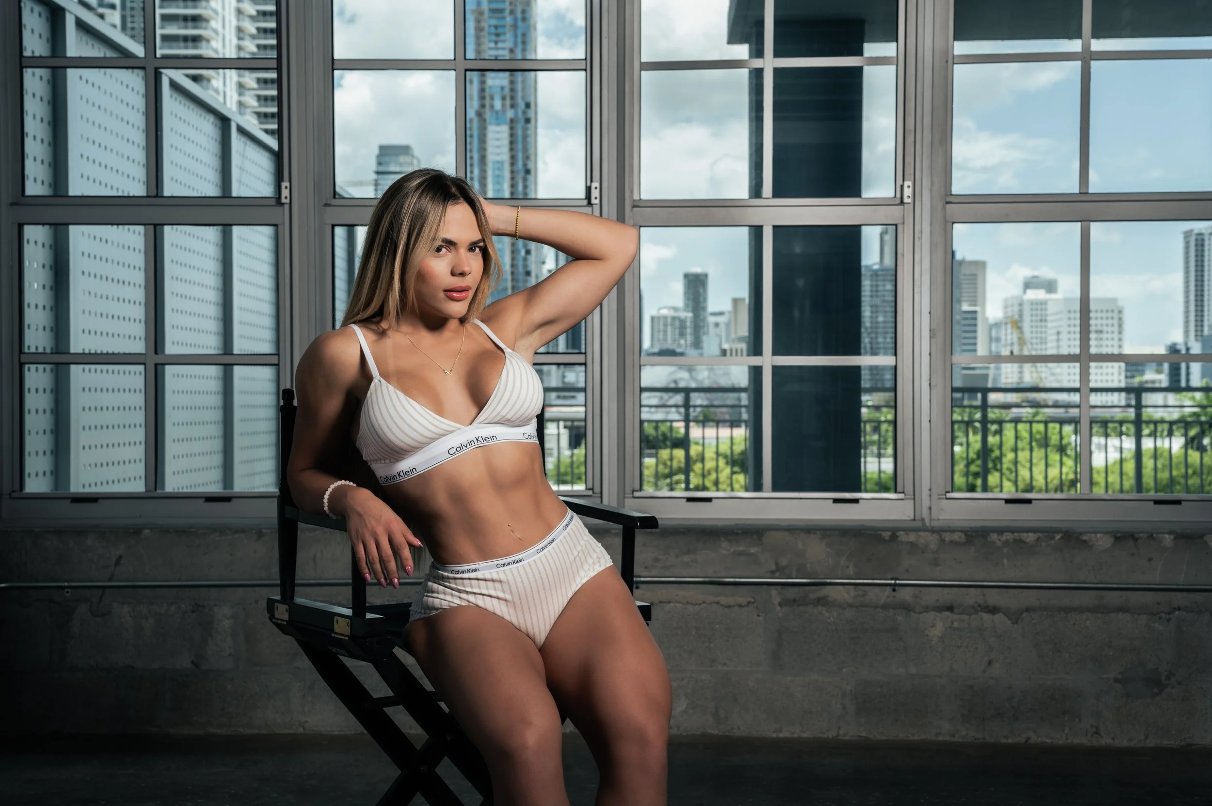 A woman with long blonde hair sitting on a black director's chair in front of large windows with a cityscape background, wearing a white Calvin Klein sports bra and matching shorts.