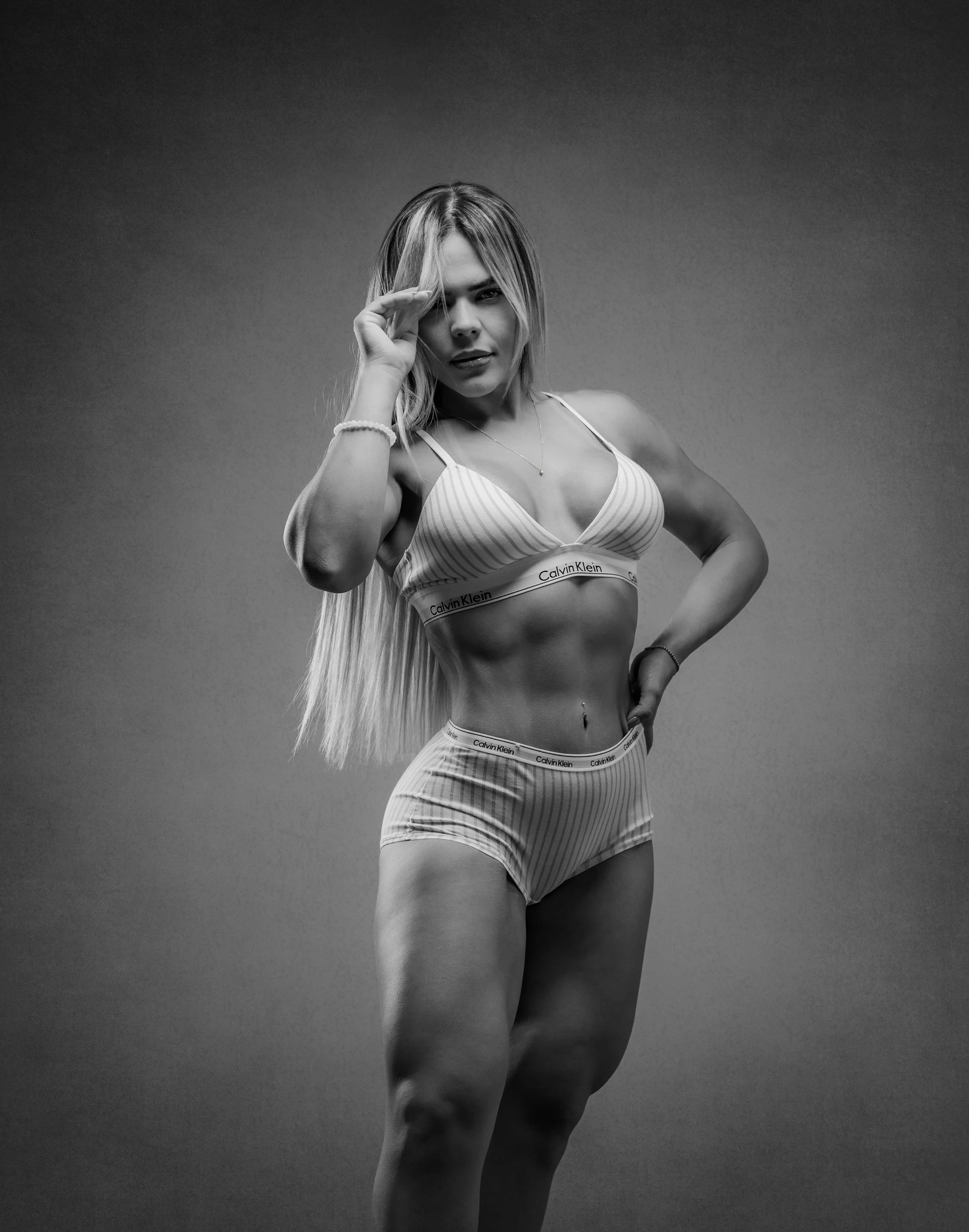 Black and white photo of a fit woman with long hair in sports bra and shorts, posing with one hand near her face and the other on her hip.