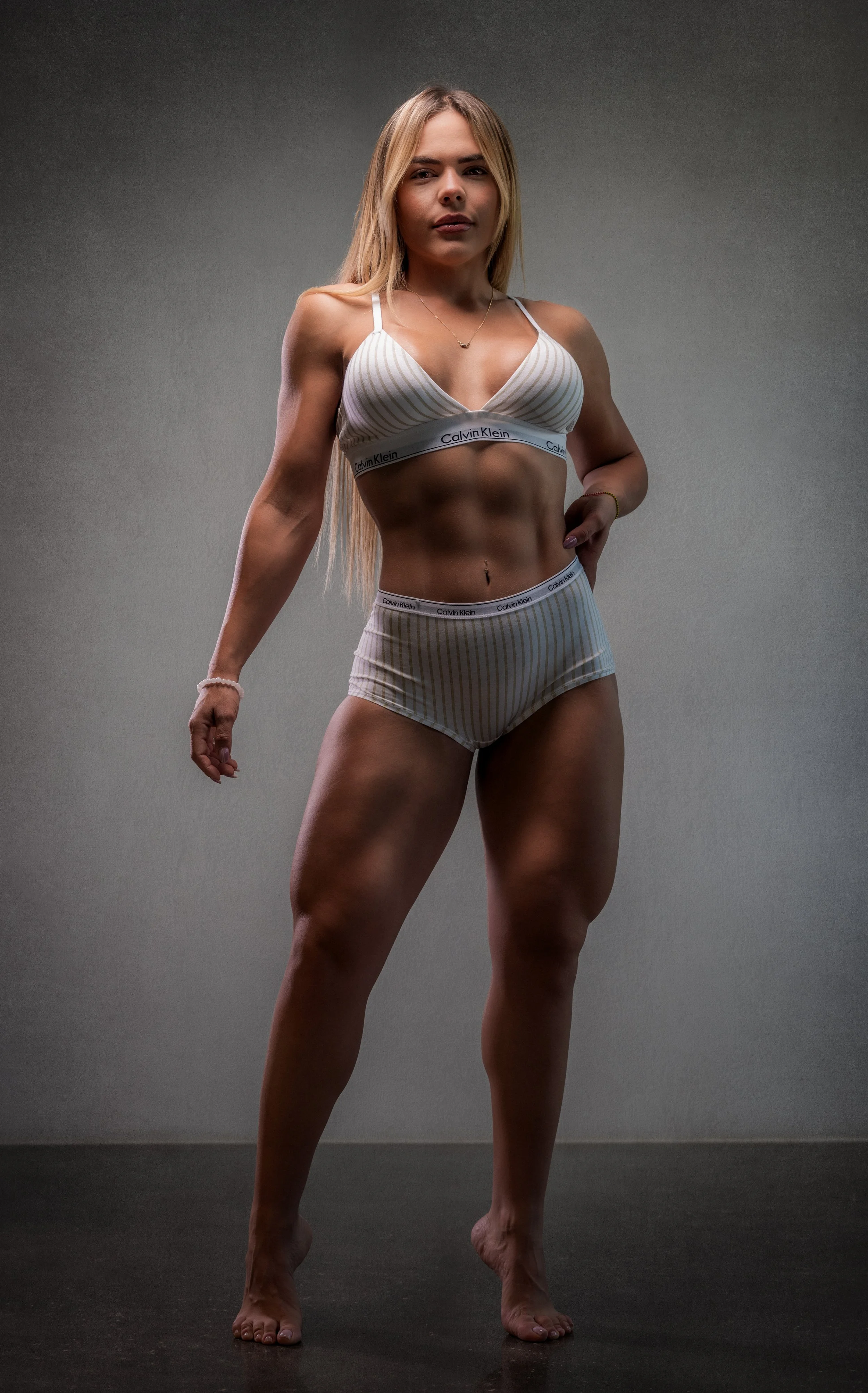 A woman with long blonde hair standing in front of a plain grey background, wearing striped Calvin Klein underwear, looking confidently at the camera.
