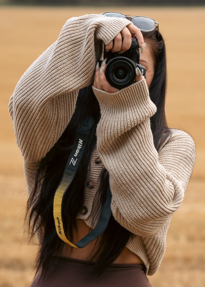 Woman with long dark hair taking a picture with a camera outdoors in a field, wearing a beige knitted sweater and sunglasses on her head.