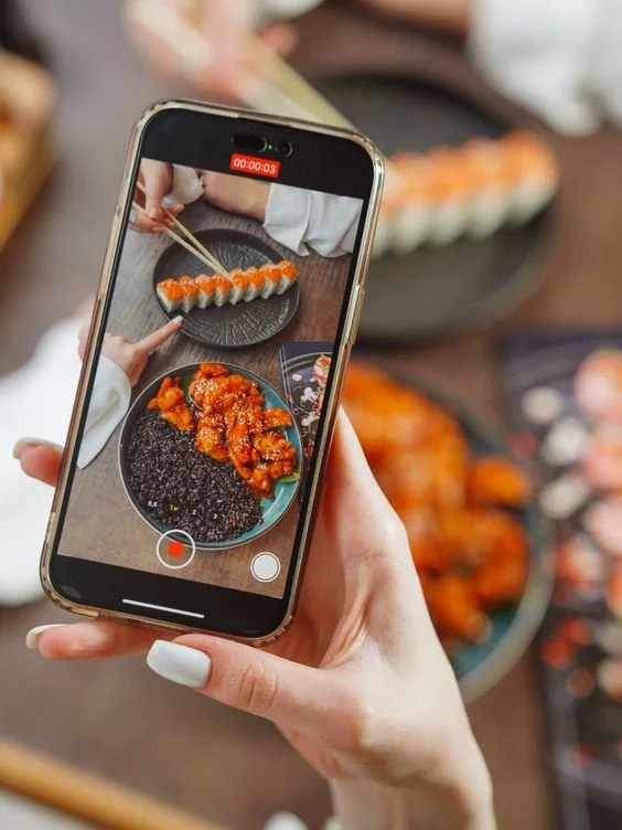 Person recording a video of a sushi platter on a smartphone, with sushi rolls and fried sushi in the background.