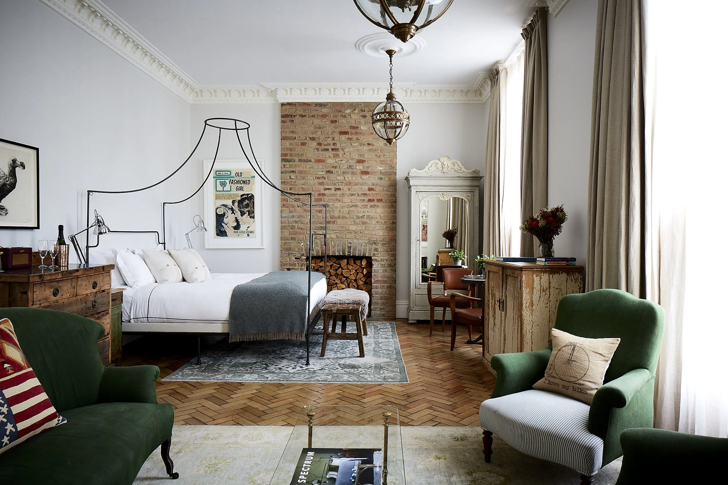 A cozy living space with a neatly made bed, a green armchair with a throw pillow, a vintage wooden dresser, and a brick fireplace with stacked firewood. The room features large windows with cream curtains, a white wardrobe with a mirror, and a small 