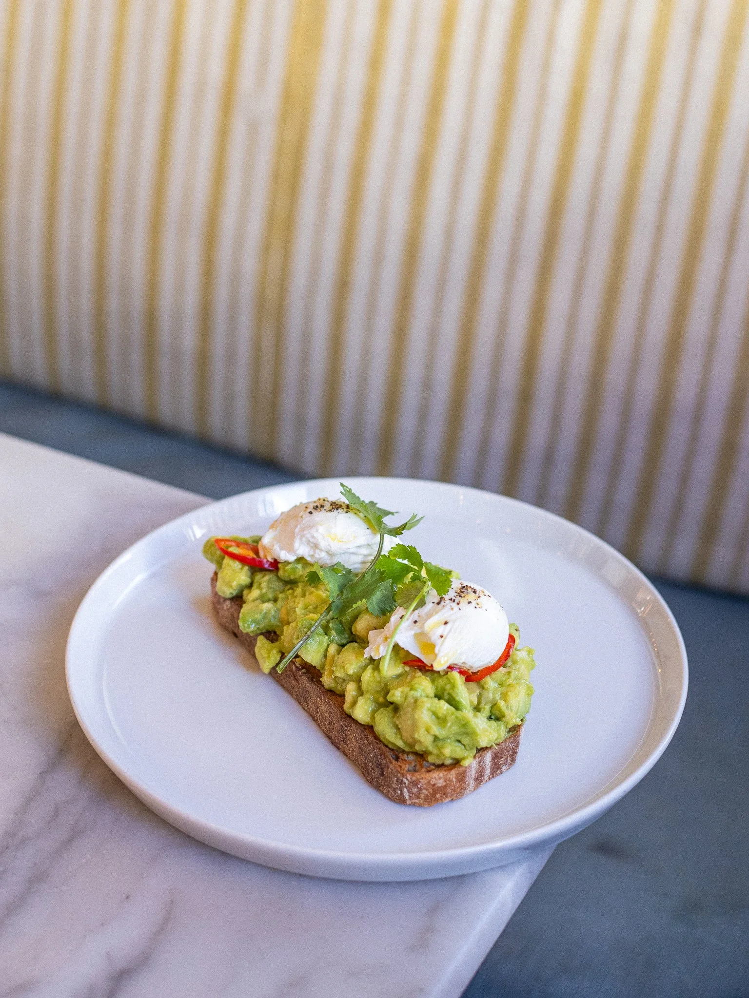Open-faced sandwich topped with mashed avocado, poached eggs, red chili slices, and cilantro on a white plate.