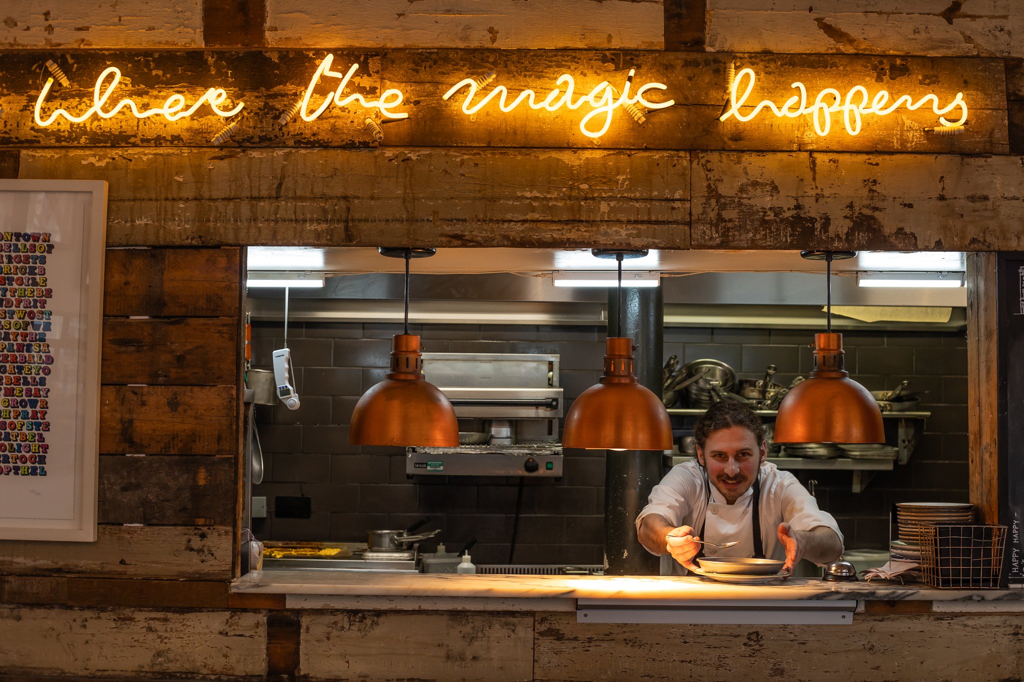 Chef smiling and reaching over a counter in a restaurant kitchen. Wooden decor and three hanging copper pendant lights. Neon sign above that reads 'where the magic happens'.