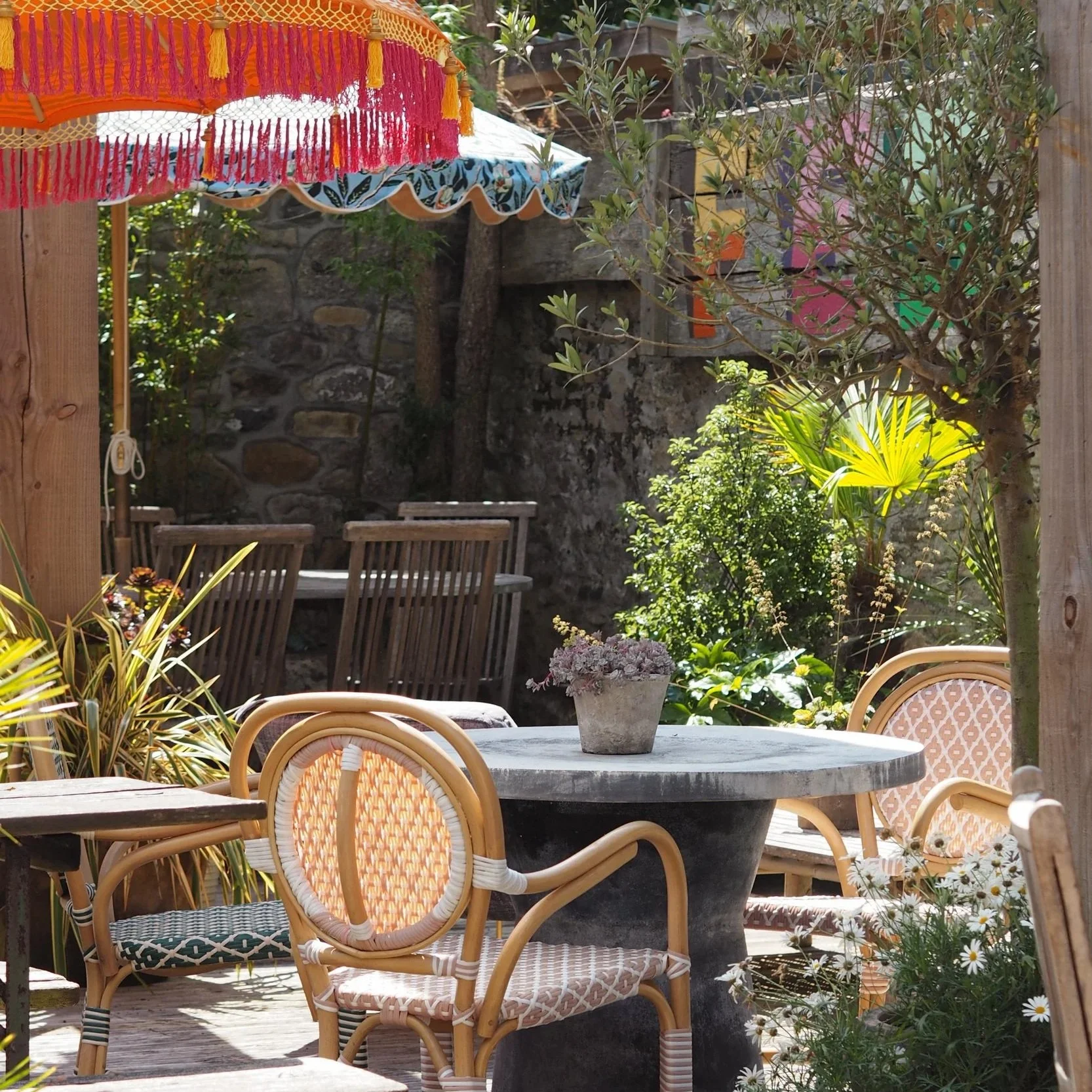 Outdoor patio area with wooden and wicker chairs, a round stone table with a potted plant, and a colorful patio umbrella, surrounded by lush greenery and a stone wall.