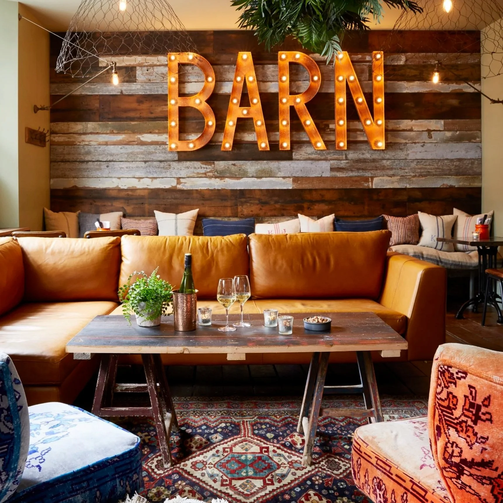 A cozy bar interior with a large illuminated 'BAR' sign on a wooden wall. Includes a mustard-colored leather sofa, a rustic wooden coffee table with wine glasses, a wine bottle, and a small potted plant. Decorative items like patterned cushions, a co
