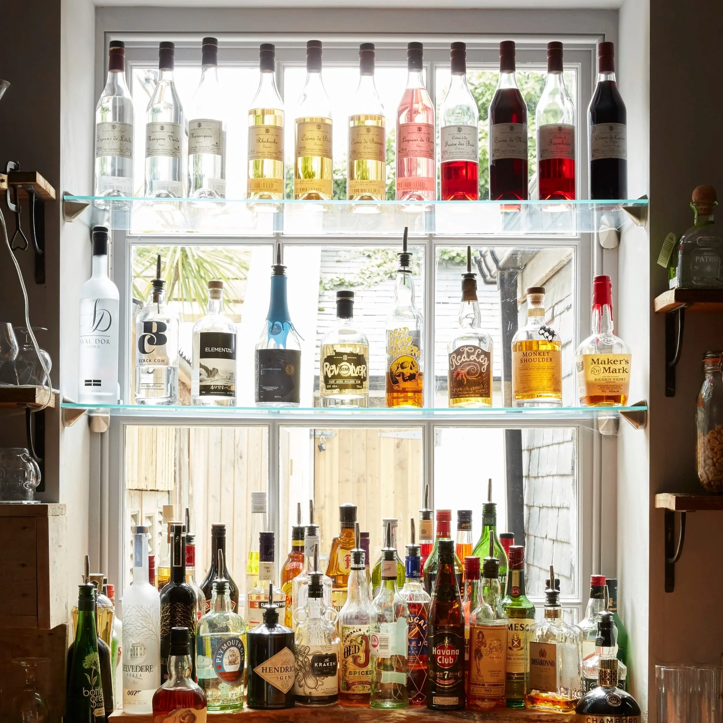 Display of various liquor bottles on glass shelves in front of a window with natural light.