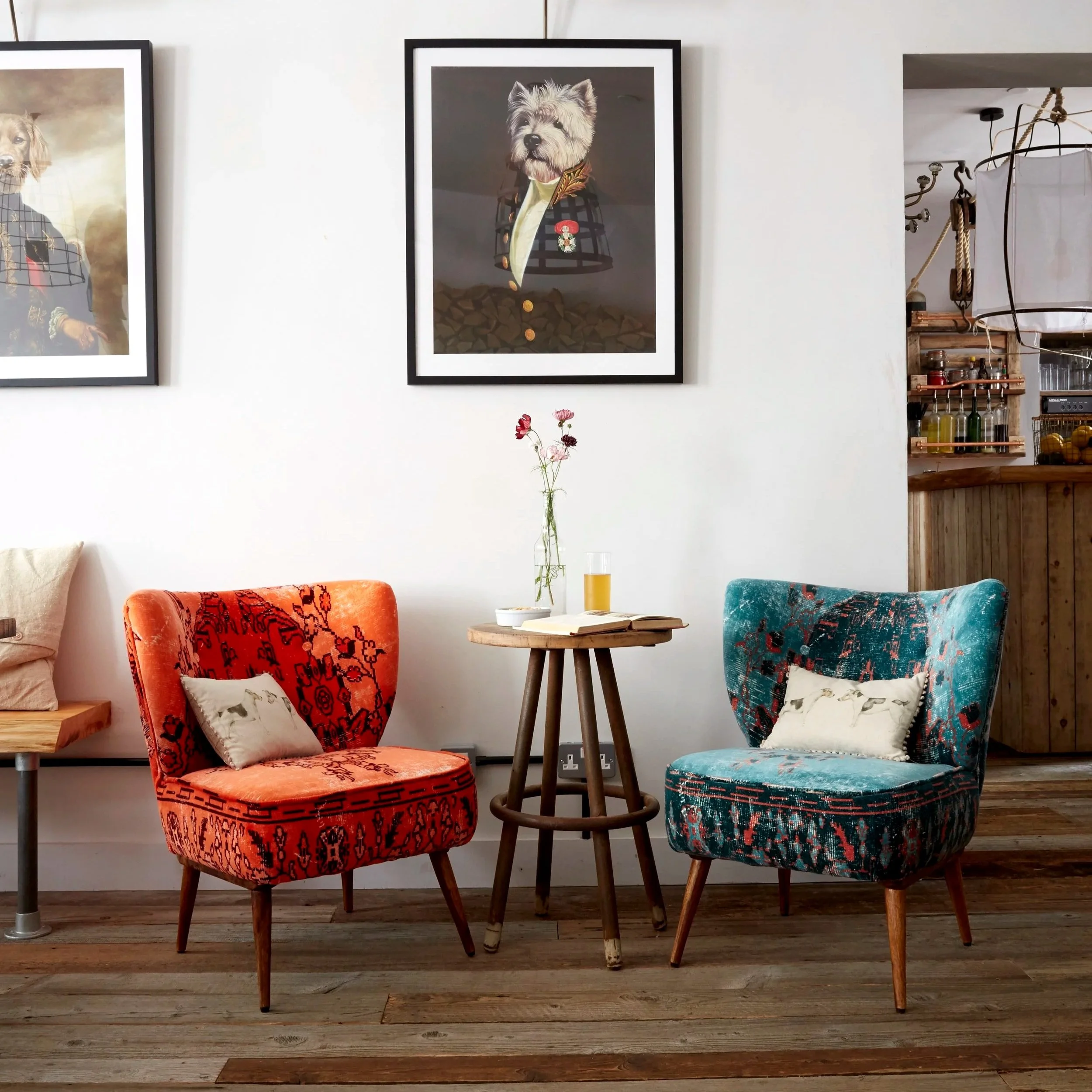 A cozy seating area with two colorful armchairs, one orange and one teal, with pillows, a small round wooden table with flowers, a drink, and books, and framed dog portraits on the white wall behind.