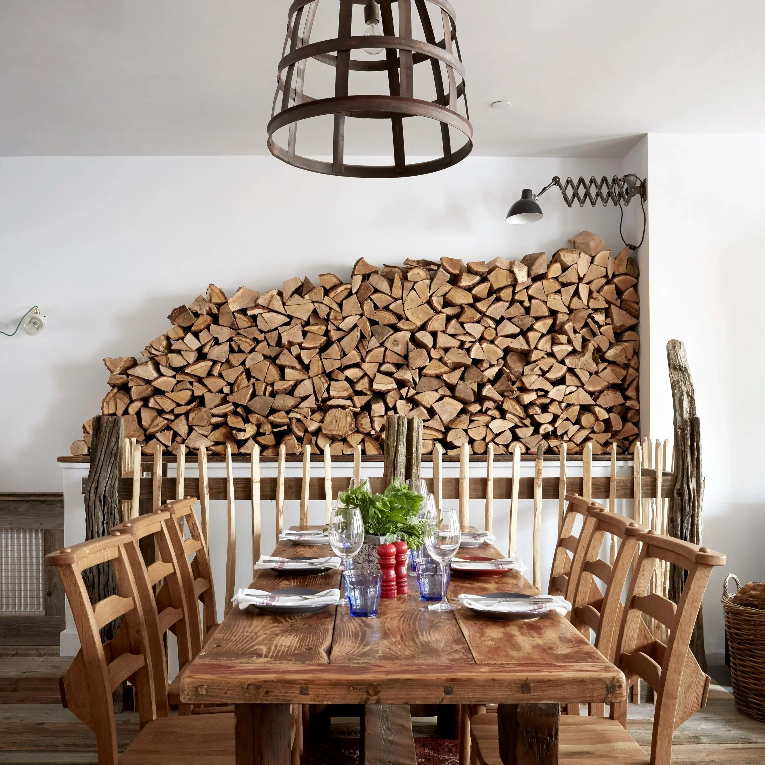 A rustic dining room featuring a wooden table with place settings, surrounded by wooden chairs, with a wall stacked with firewood and a wooden fence at the back, adorned with natural wooden logs.