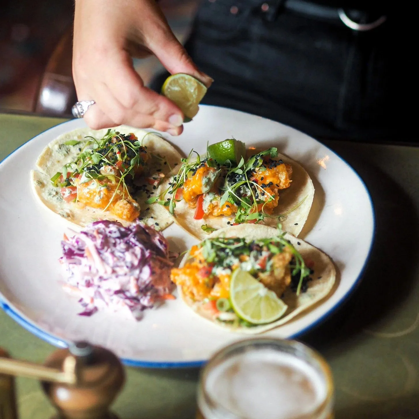 A white plate with four fish tacos topped with greenery and cilantro, a serving of coleslaw, and a lime wedge, with a hand squeezing lime over the tacos, and a glass of beer in the foreground.