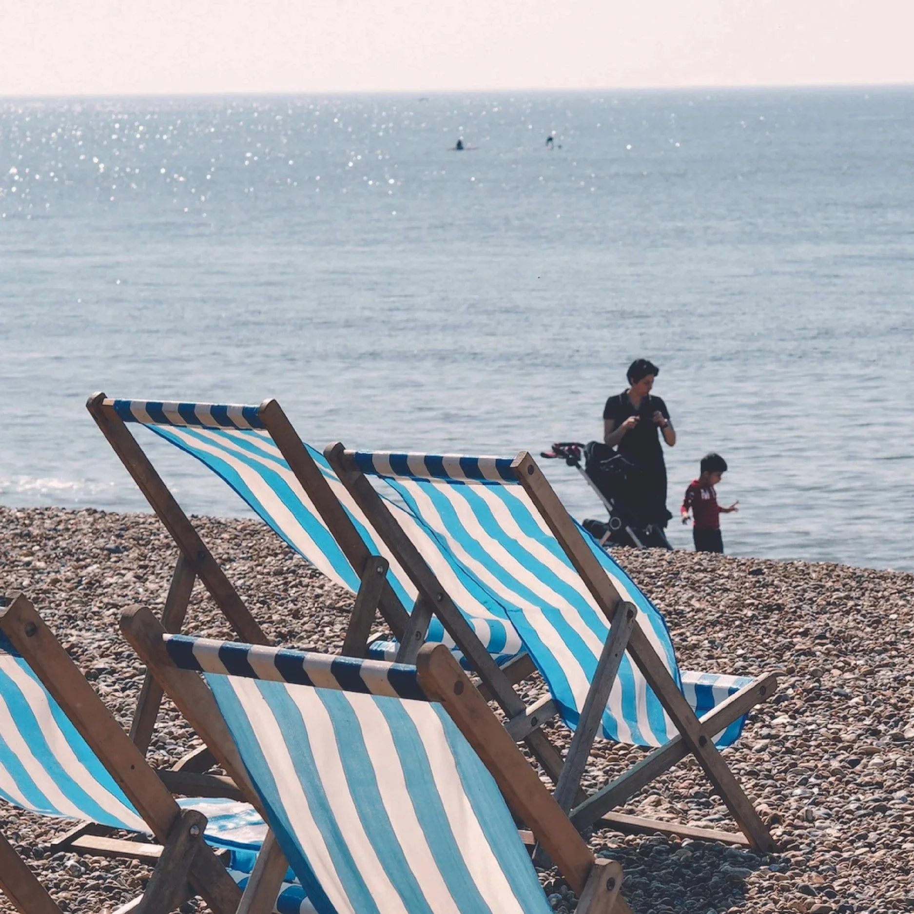 Row of blue and white striped deck chairs on a pebbled beach with people in the water and a clear sky in the background.