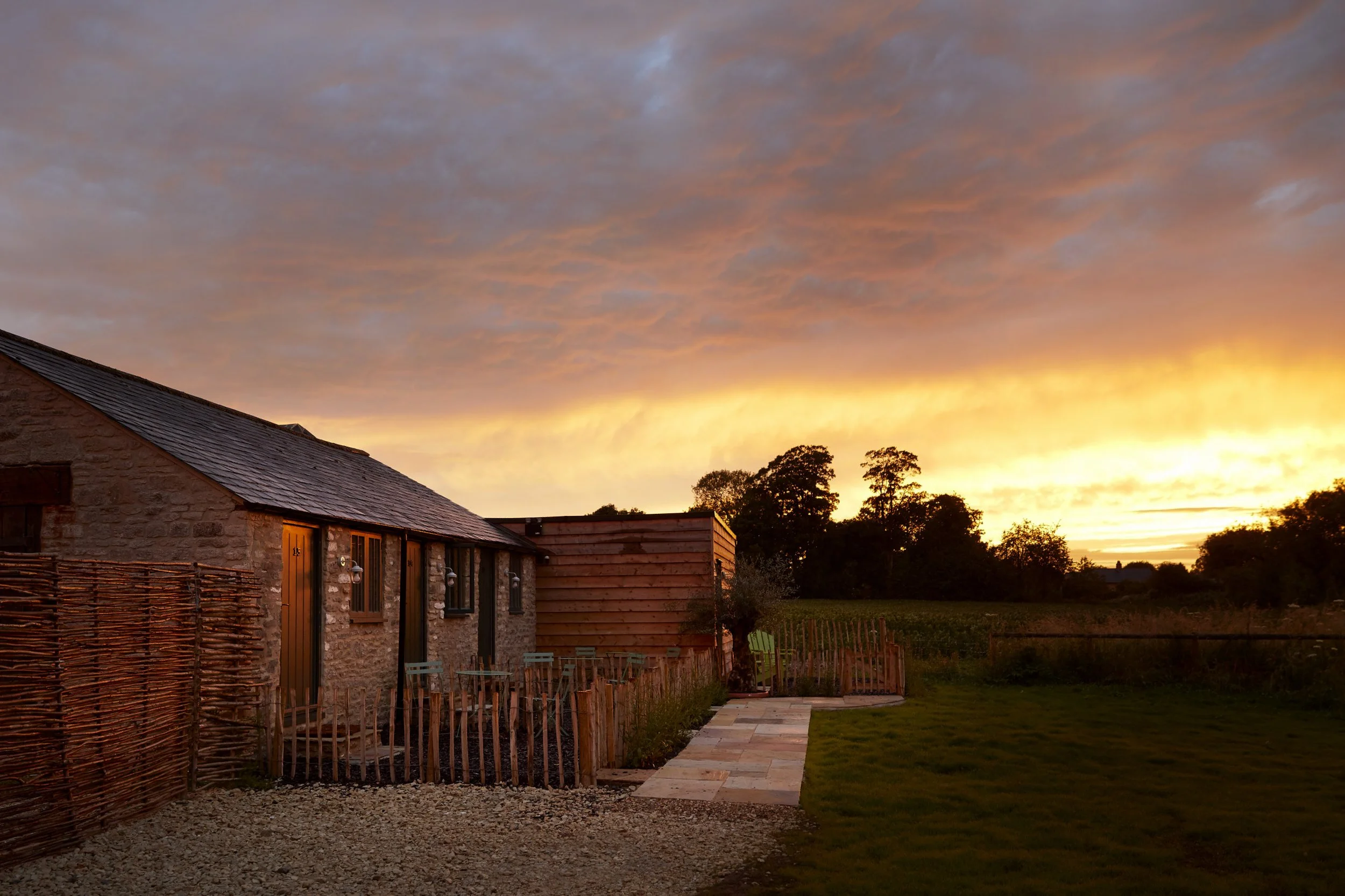 A rustic stone house with a slate roof during sunset, with a small garden area and a pathway leading to a wooden fence, surrounded by trees and open fields.