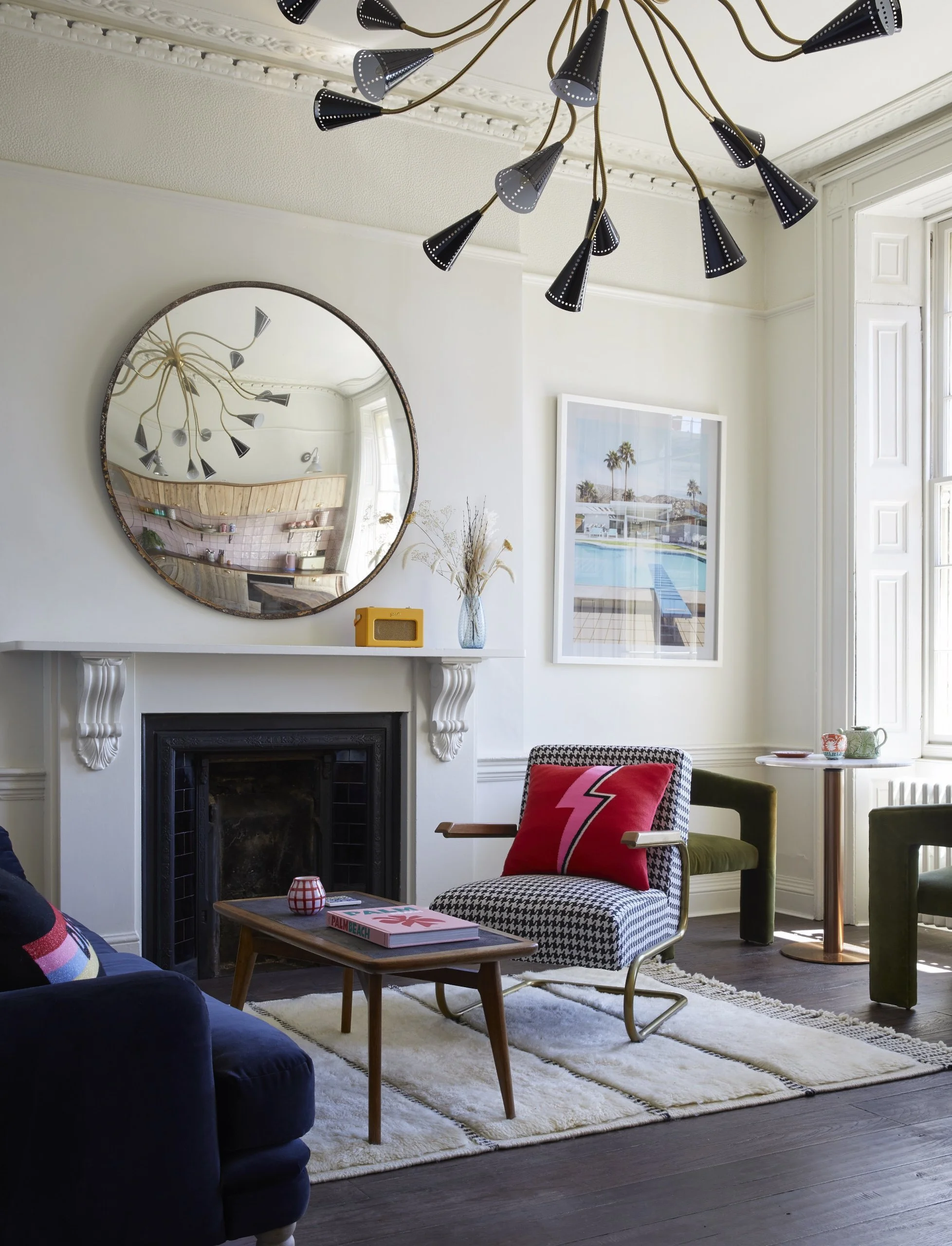 Living room with a white fireplace mantel, a round mirror, a black and white patterned chair with a pink lightning bolt pillow, a small wooden coffee table, a navy blue sofa, a framed photo of a pool scene, a round side table with cups, and a modern 