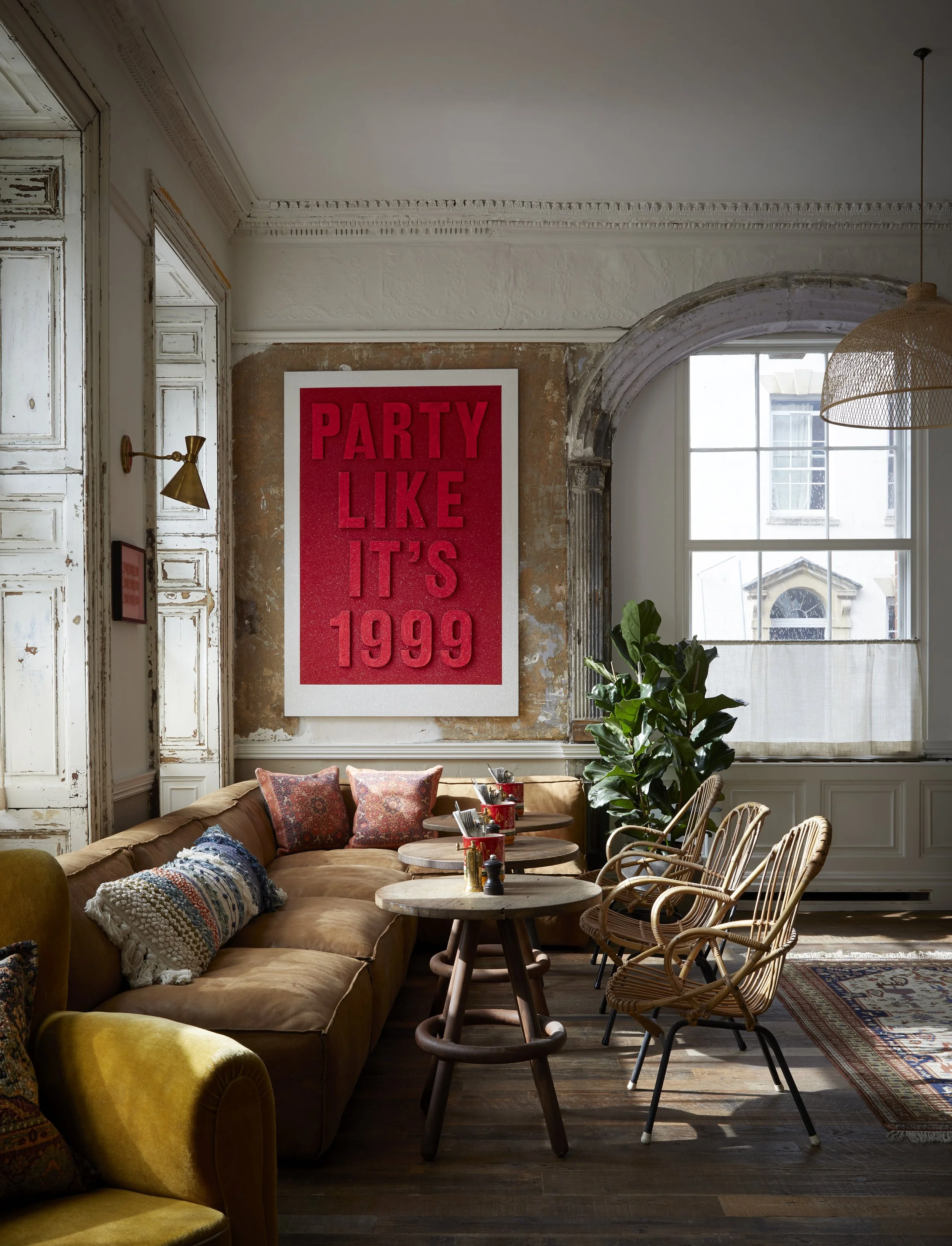 Interior of a stylish living room with a large beige sectional sofa decorated with colorful pillows, surrounded by round wooden tables, bamboo chairs, and a large window with white curtains. A prominent red wall art reads 'PARTY LIKE IT'S 1999,' with a tall green plant nearby.