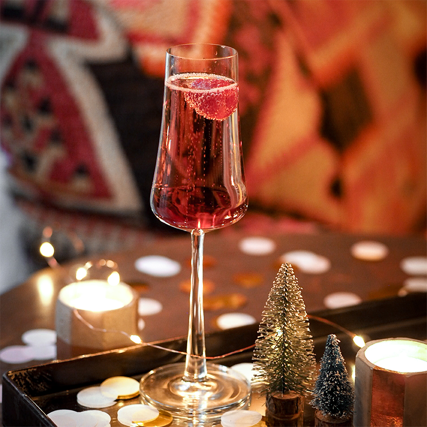 A tall glass of pink sparkling beverage with a slice of strawberry inside, placed on a table decorated with small fairy lights, miniature evergreen trees, and scattered white and gold confetti, with a lit candle in the background.