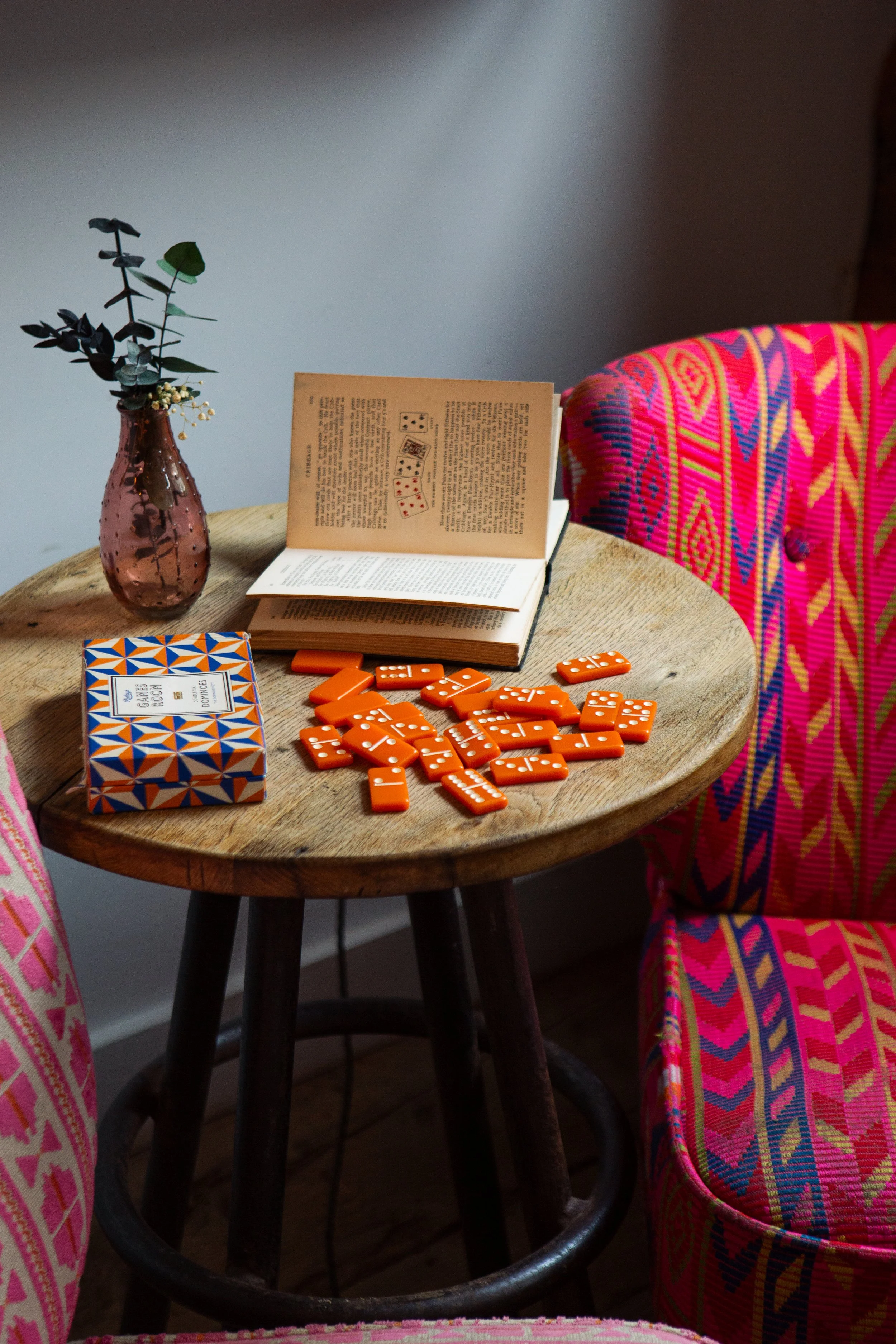 A round wooden table with a vase of greenery, an open book, orange domino pieces, a deck of card game, and two colorful patterned chairs.