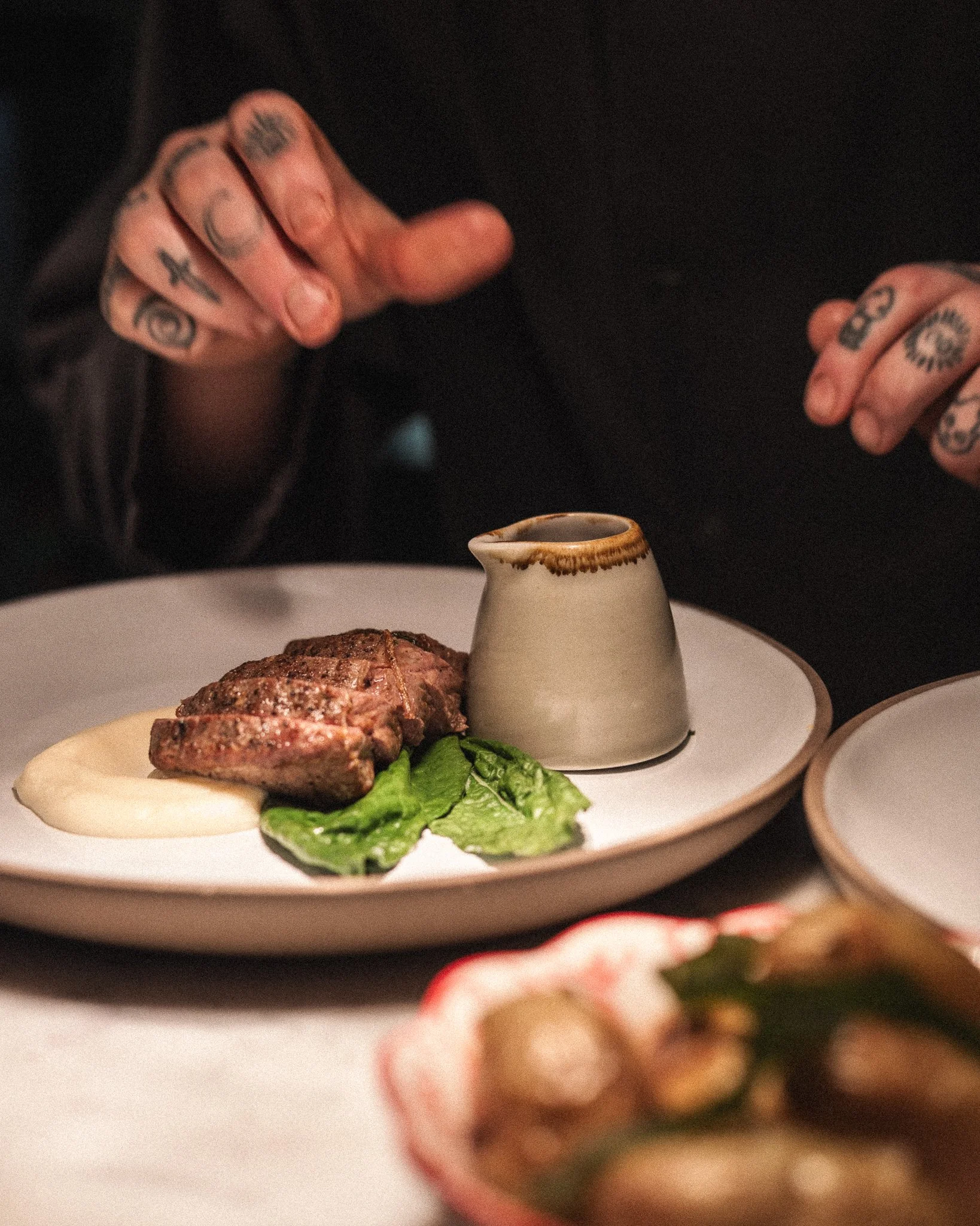 Close-up of a plate with grilled meat, fresh greens, a small ceramic pitcher, and a person with tattoos preparing to serve