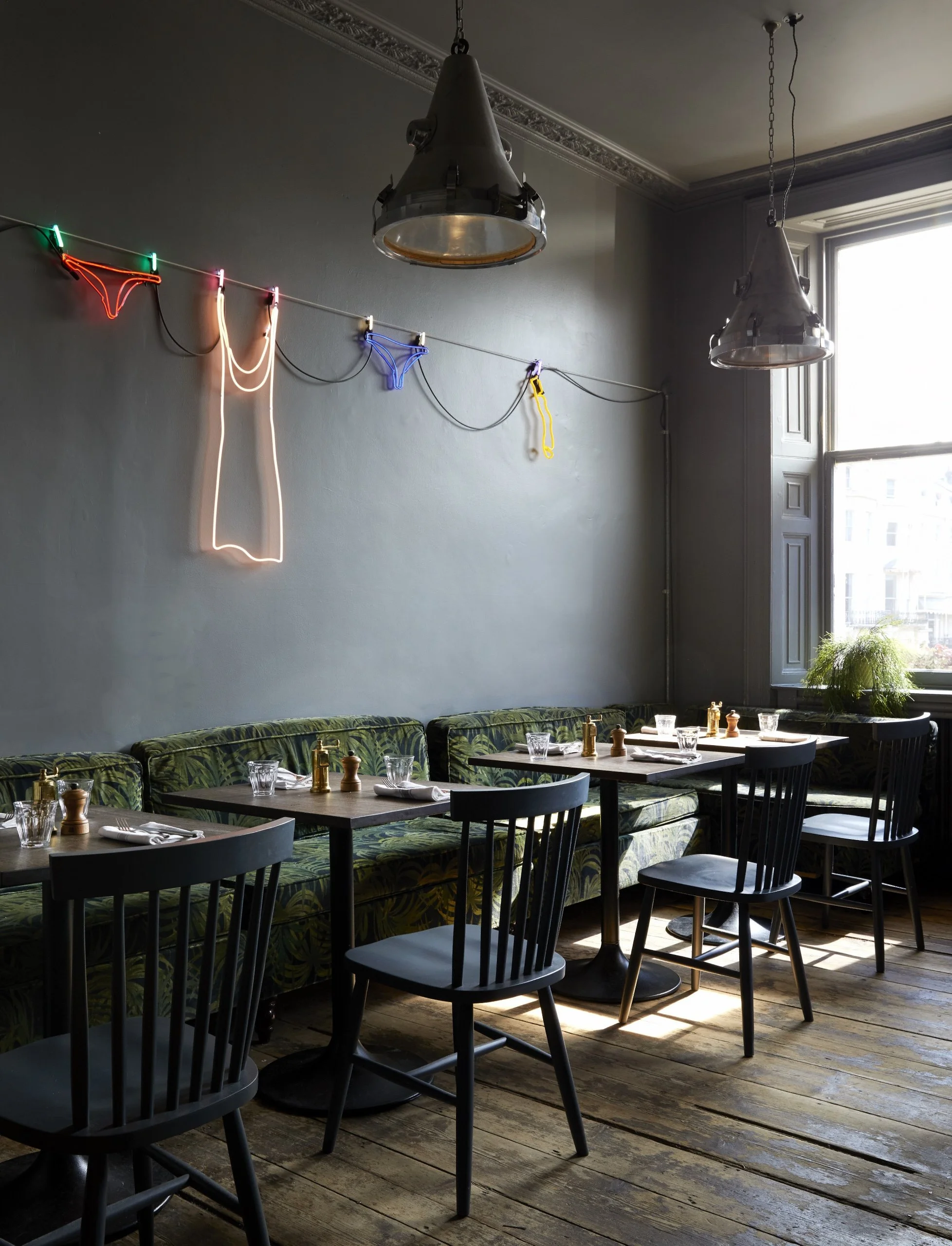 A cozy indoor restaurant with wooden floors, a row of tables set with glasses, salt and pepper shakers, and napkins. There are black chairs and a patterned cushioned bench along the wall. Overhead, modern metallic pendant lights and a neon sign of a party girl silhouette are visible, with natural light coming in from a large window.