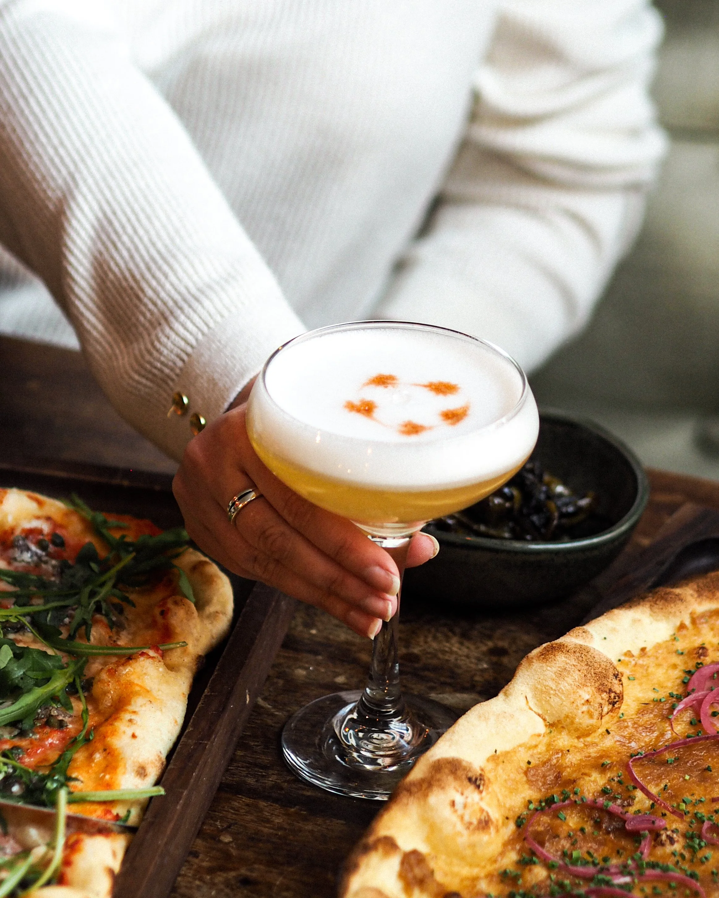 Person holding a cocktail glass with foam and orange garnishes, surrounded by pizzas on a wooden table.