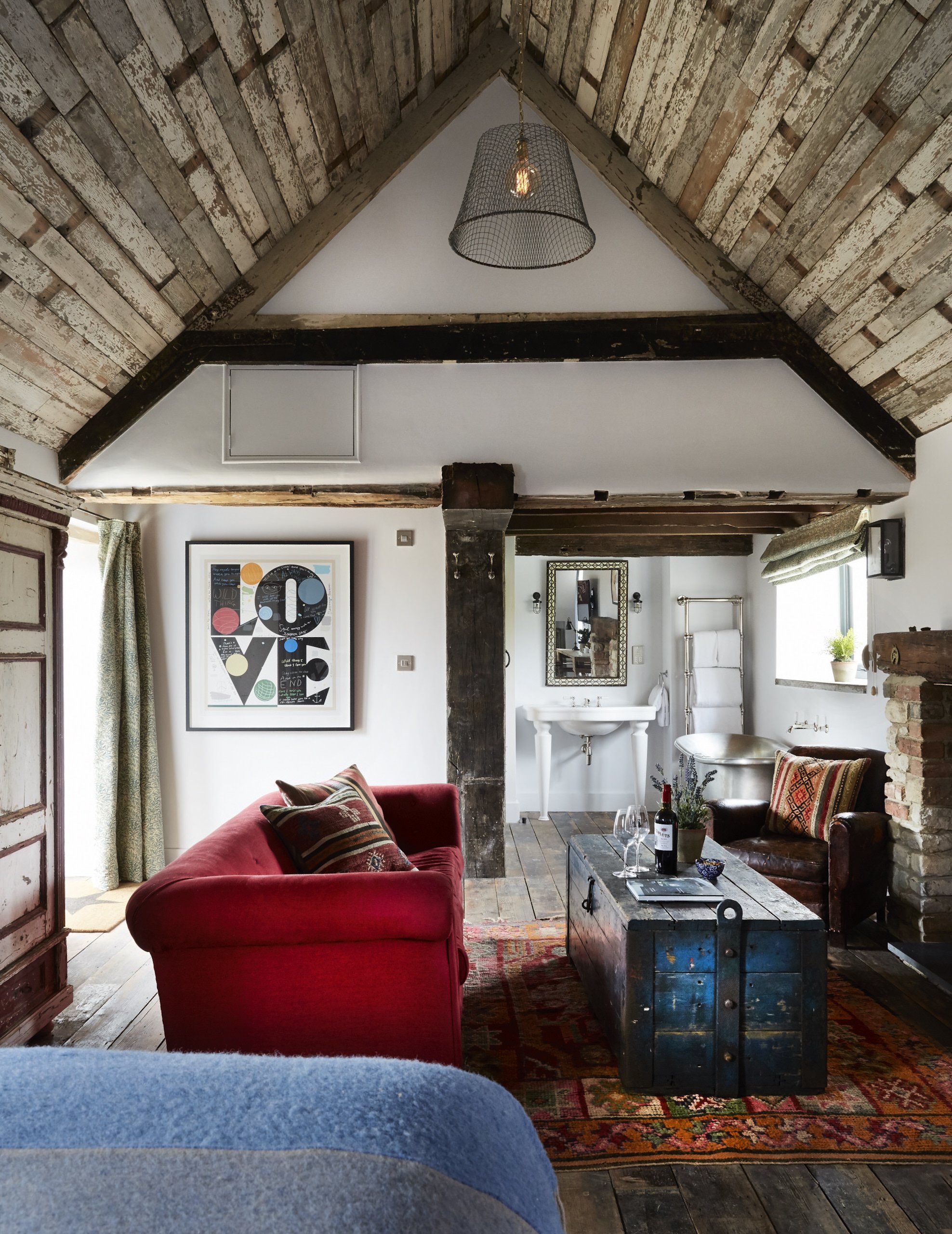 Cozy living room with a red sofa, patterned pillows, rustic wooden ceiling, vintage trunk coffee table, and exposed brick and wood accents, with a small bathroom visible in the background.