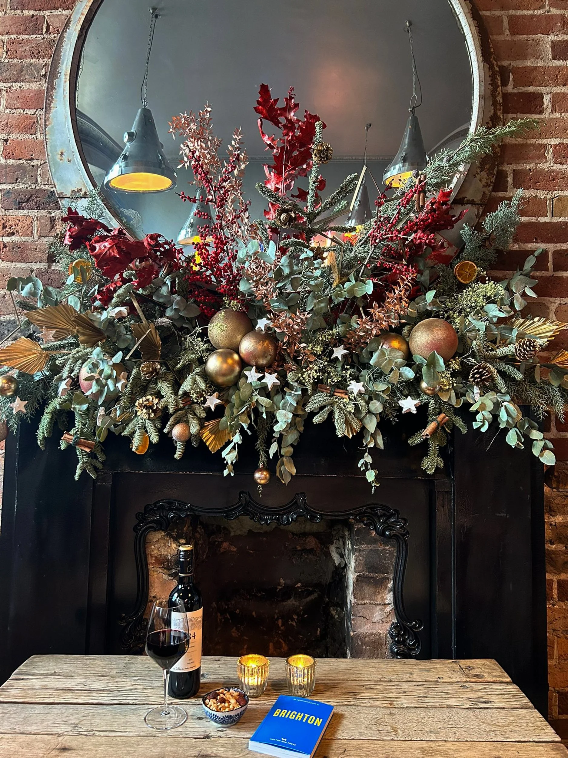 A festive table setup with a bottle of red wine, a glass of red wine, a bowl of nuts, two lit candles, a book titled 'Brighton', and a small bowl of snack mix, with a decorative arrangement of greenery, berries, ornaments, and dried citrus on a fireplace mantel in the background.