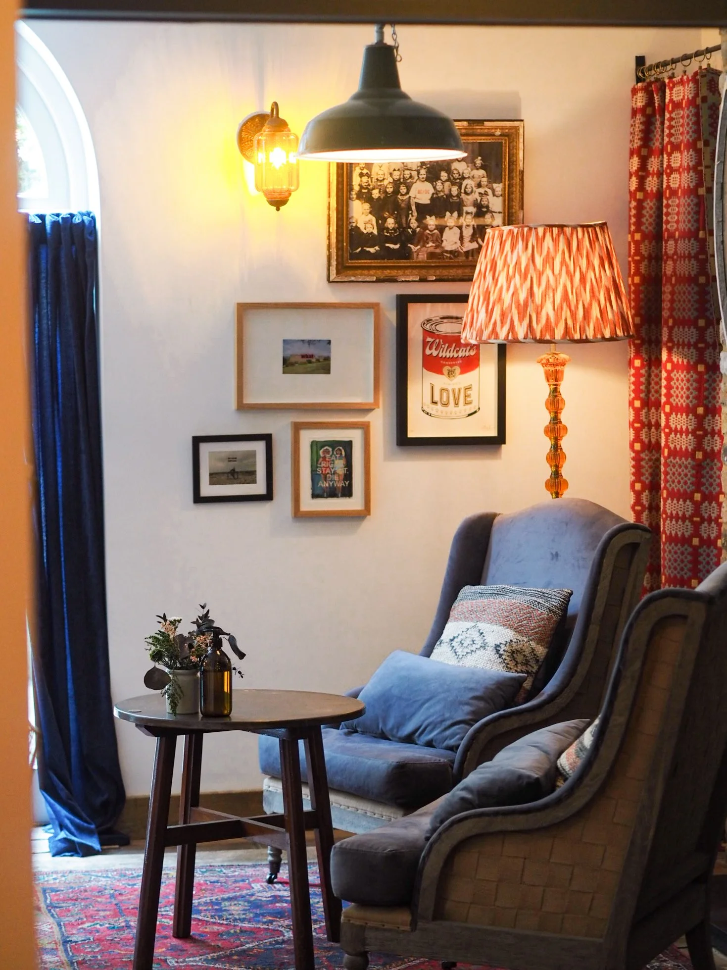 A cozy living room corner with a gray armchair that has a decorative pillow, a round wooden side table with a woven vase containing flowers, a vibrant red and blue patterned rug, a framed family photo collage, a vintage-style lamp, and curtains with a red and white geometric pattern.
