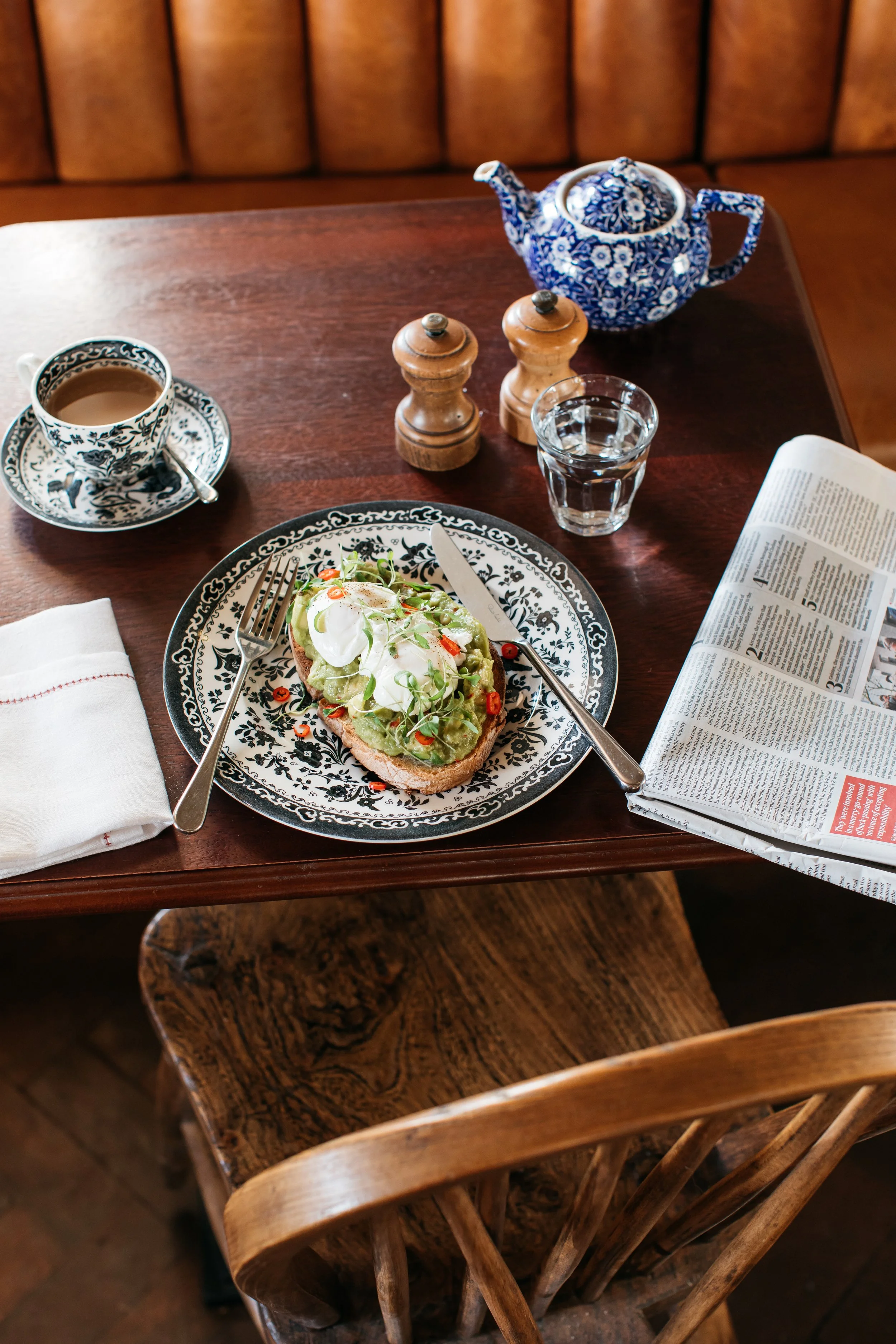 A wooden table in a cozy cafe with a patterned teapot, a cup of tea, a glass of water, a plate with toast topped with avocado, poached eggs, and microgreens, salt and pepper shakers, an open newspaper, and a white napkin.