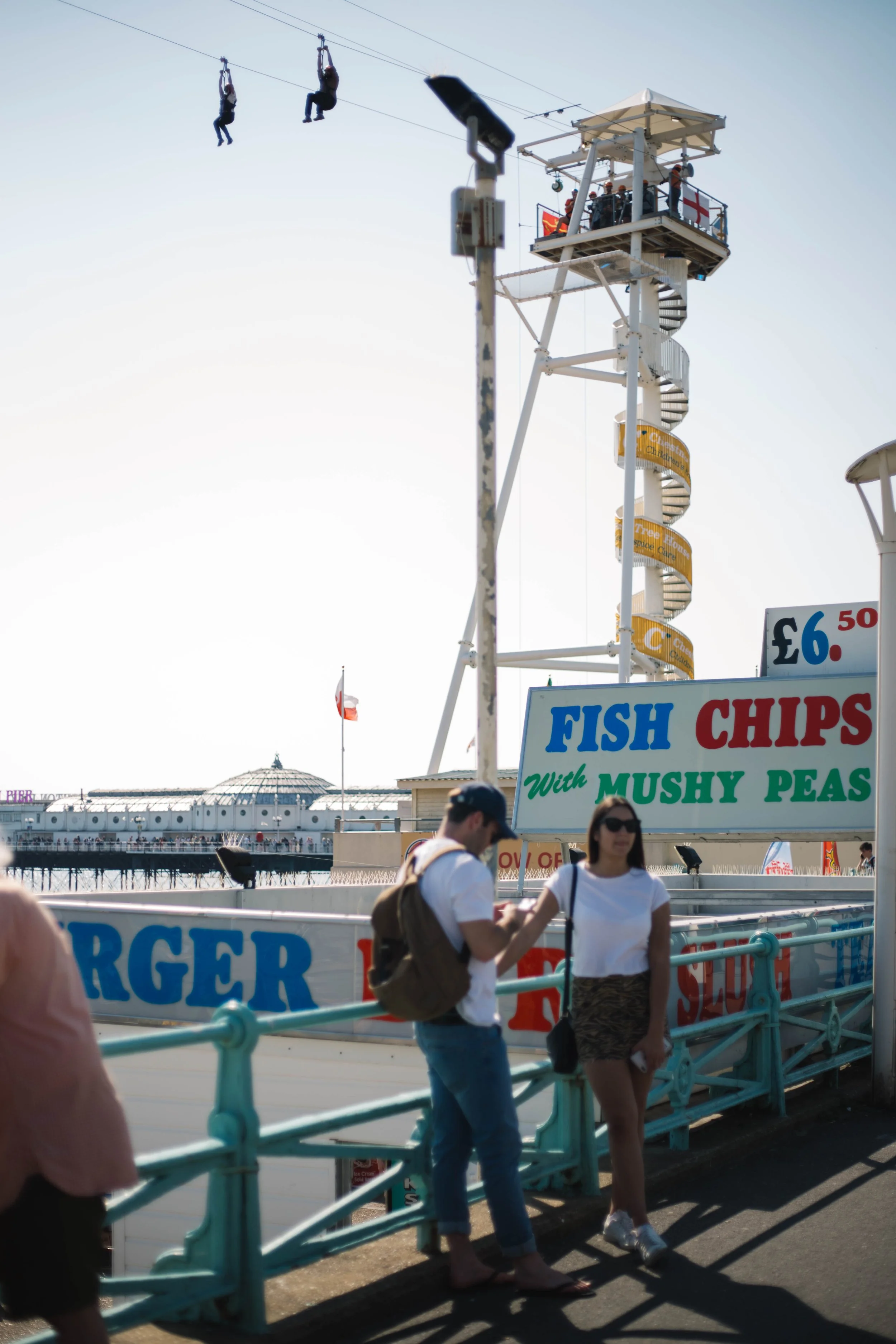 People walking on a pier with a sign advertising fish and chips with mushy peas, and a large amusement ride in the background, including a Ferris wheel and a tower with spiral stairs, near the coastline.