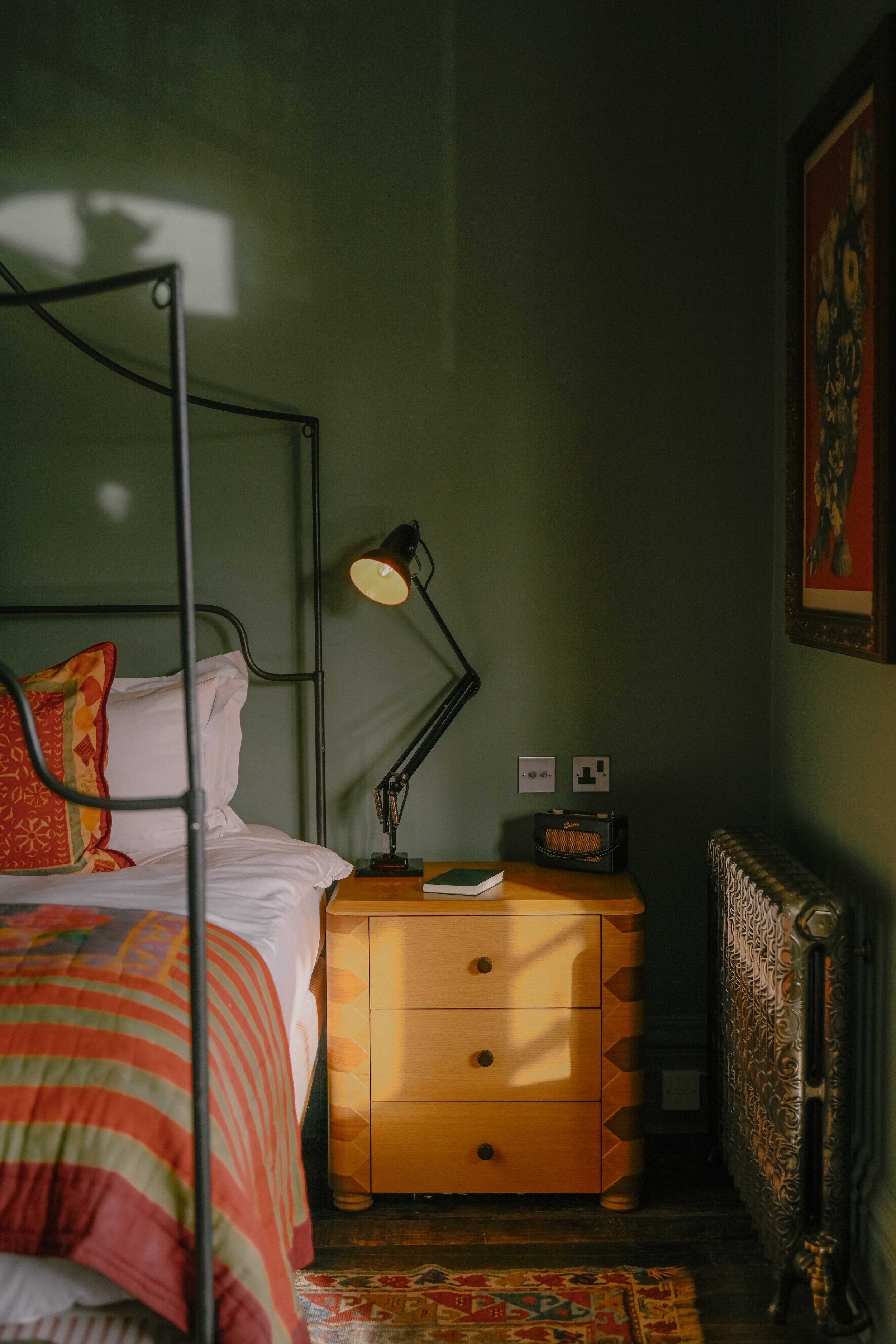 A bedroom with a green wall, a bed with white and colorful bedding, a nightstand with a book and a lamp, a framed artwork on the wall, and a radiator.