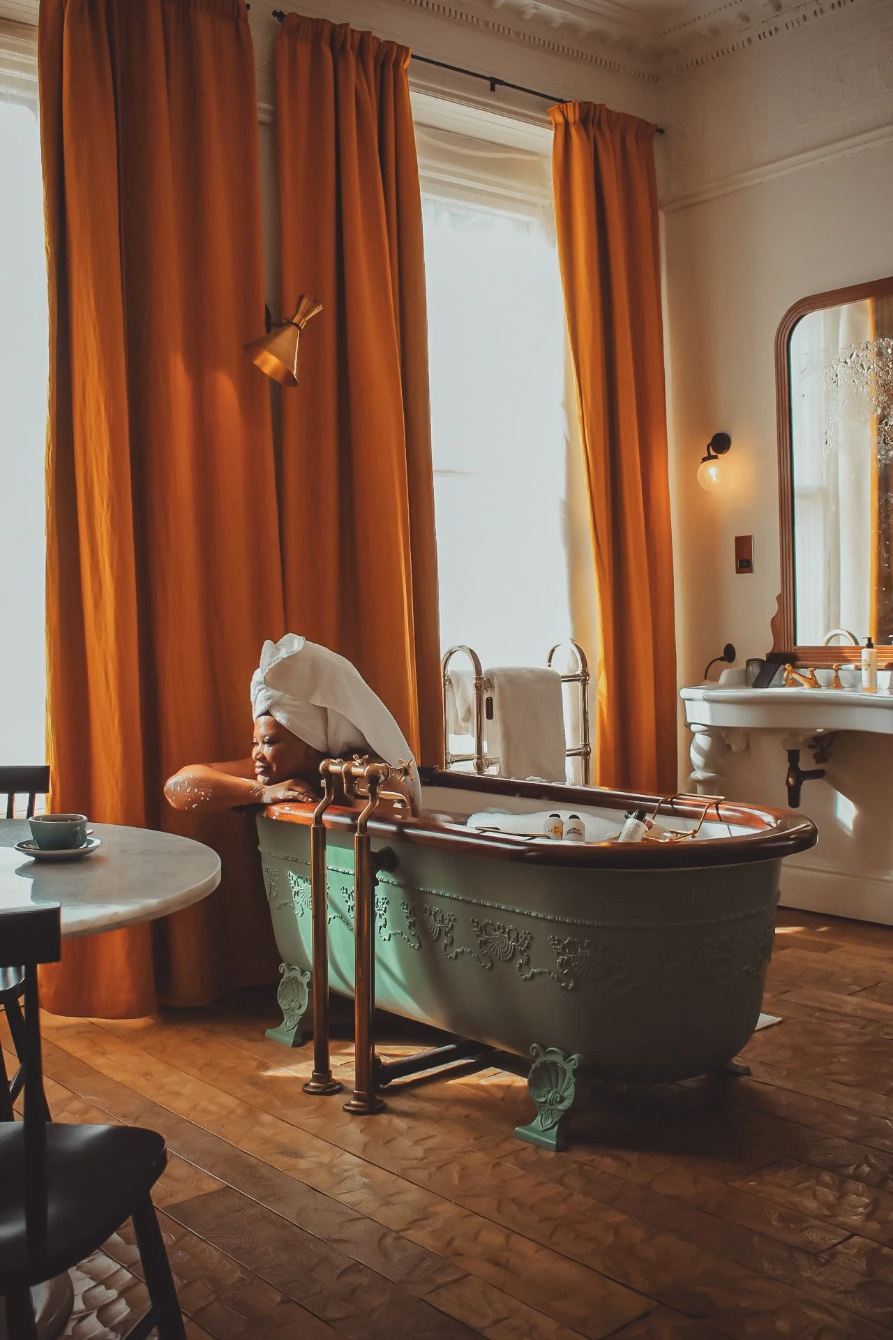 A woman relaxing in a vintage bathtub in a warmly lit room with large amber curtains, a round marble table, and a white sink.