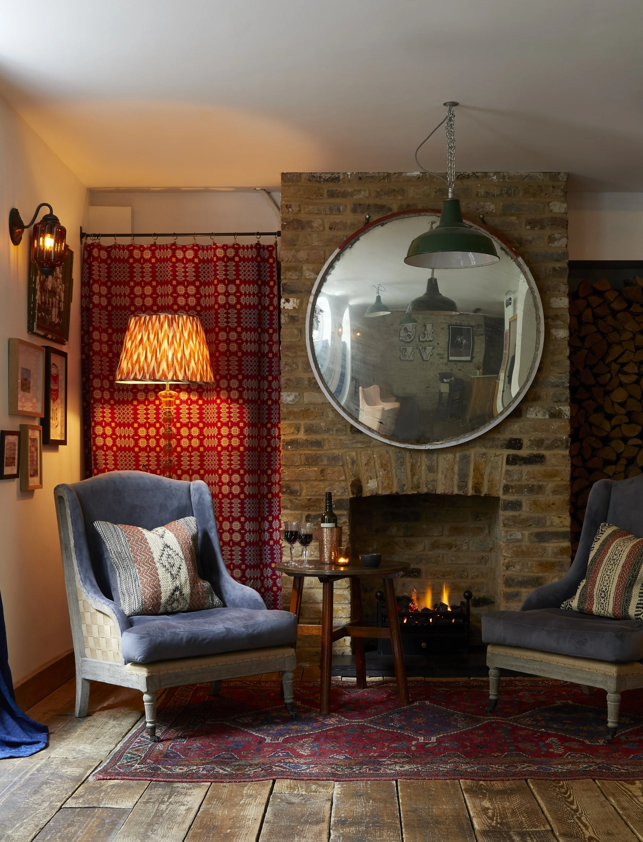 Cozy living room with two gray upholstered armchairs, a small round wooden table with wine and candles, a brick fireplace with a small fire, a large circular mirror on the brick wall, a red patterned curtain, a red and black patterned rug, and a yellow lamp with a patterned shade. A stack of firewood is visible on the right side.