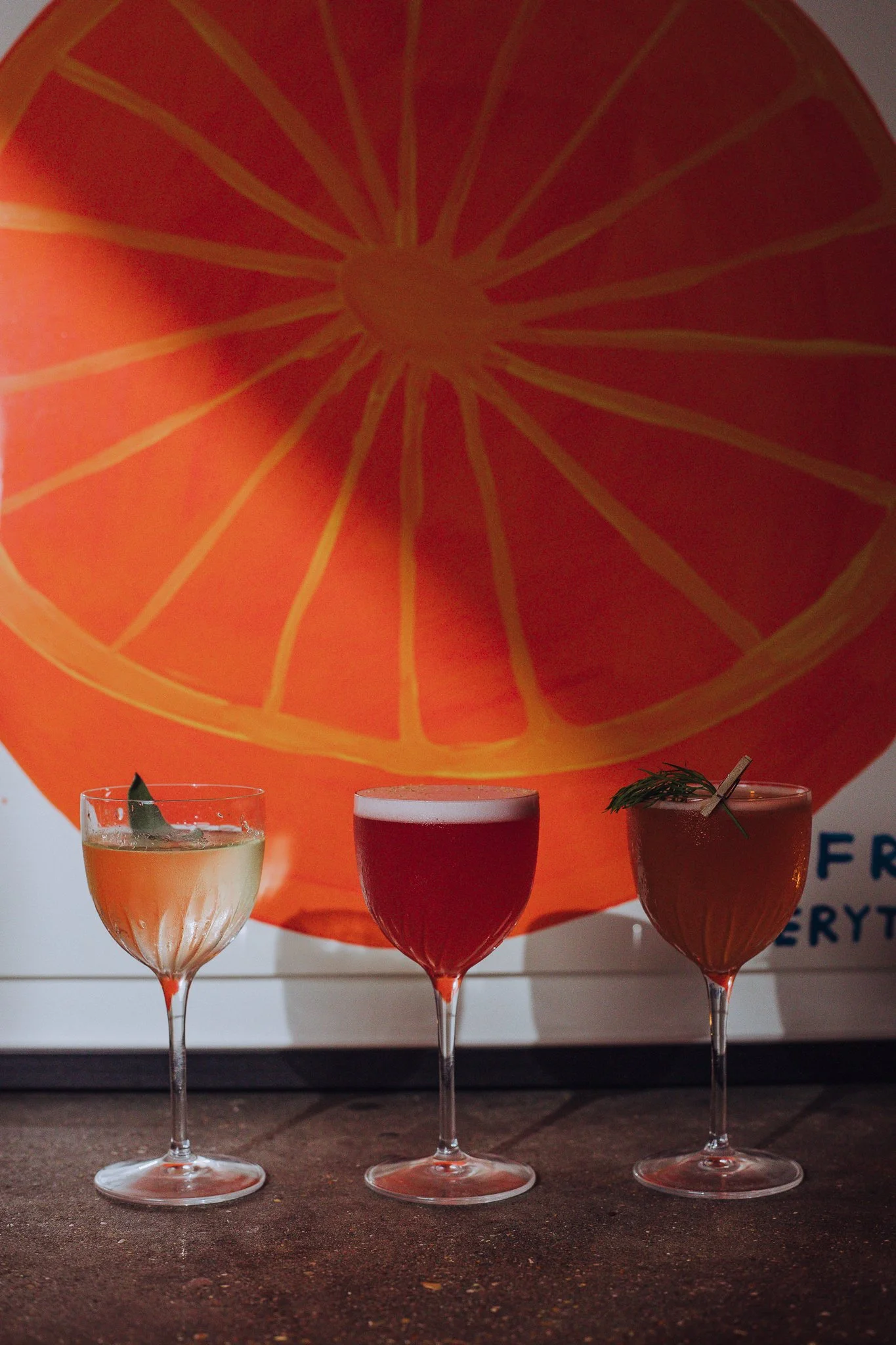 Three cocktail glasses on a table in front of a large poster of an orange slice. The drinks include a yellowish cocktail with a leaf garnish, an amber beer, and a reddish-brown cocktail garnished with herbs.