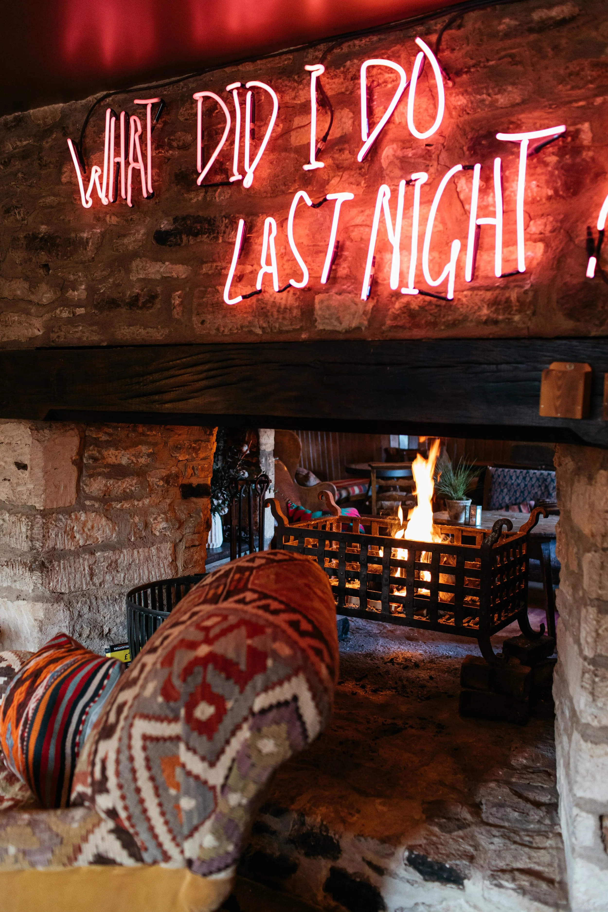 A cozy indoor scene with a stone fireplace, a fire burning inside a black metal grate, a wooden mantel above, and a neon sign that reads 'What Did I Do Last Night?'. The image includes a patterned fabric-covered object in the foreground.