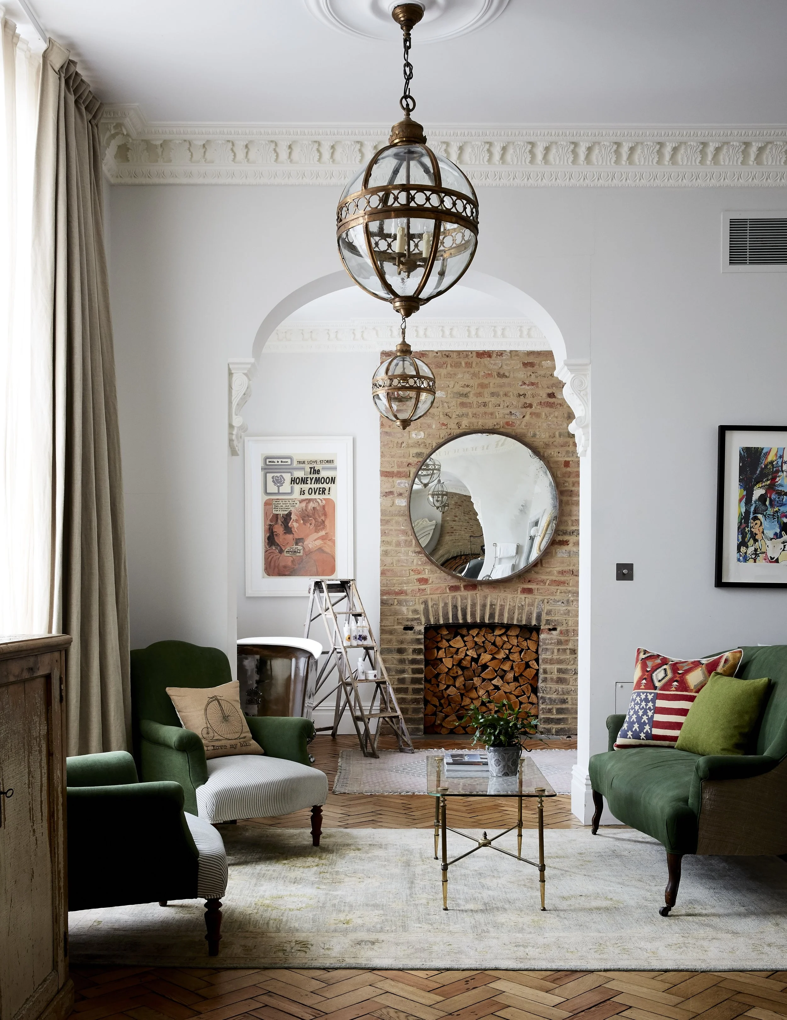 A living room with white walls, featuring a brick fireplace with a stack of logs inside and a large round mirror above it. The room has two green velvet armchairs, a beige armchair with a bicycle print pillow, a glass coffee table with a potted plant, and artwork on the walls. There are two ornate hanging light fixtures, a ladder, and a curtain on the side. The furnishing and decor are cozy and eclectic.