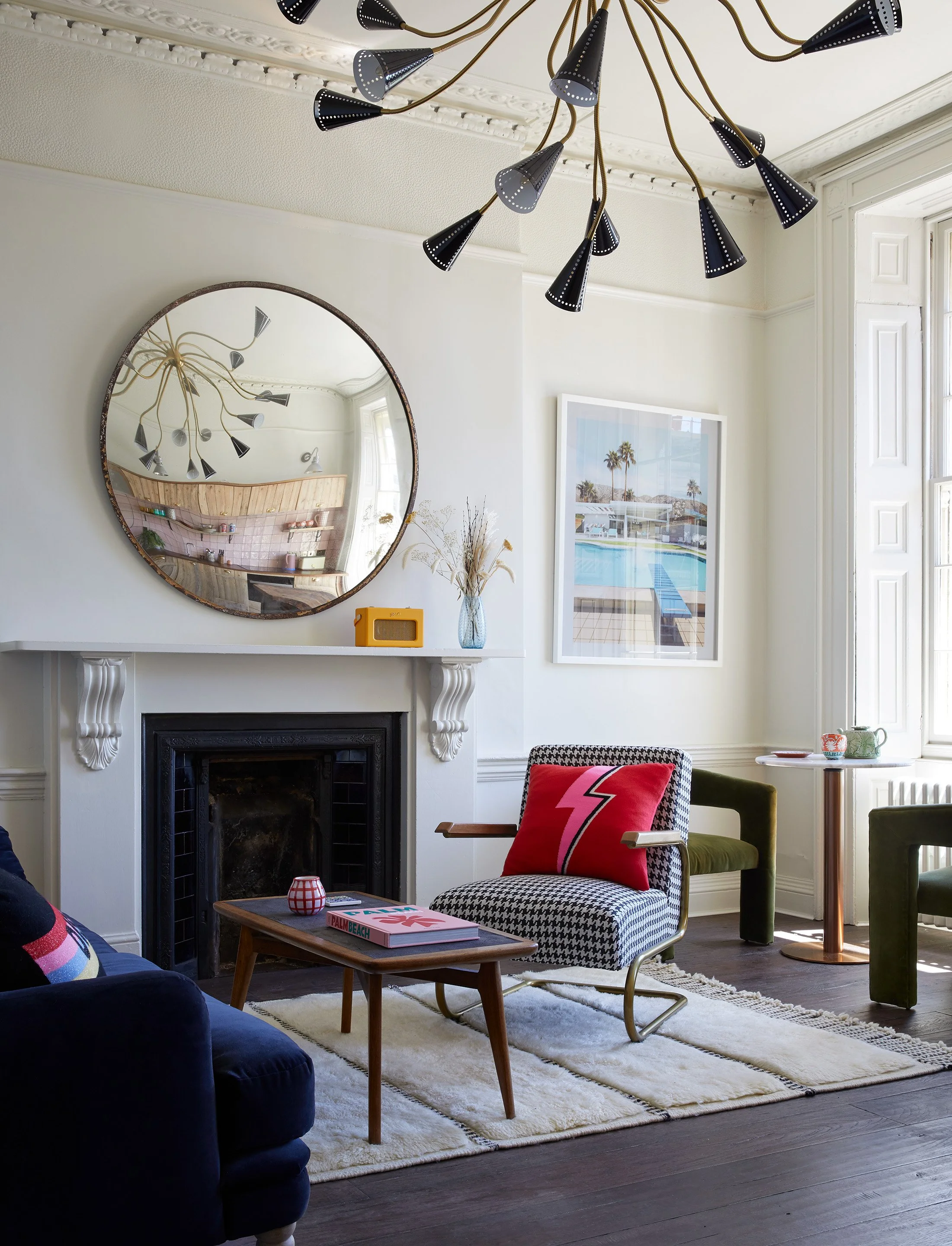 A cozy living room with a fireplace, a wooden coffee table, and armchairs, including one with a lightning bolt pillow, a white rug, a round wall mirror, a modern ceiling light, and wall art featuring palm trees with a pool.