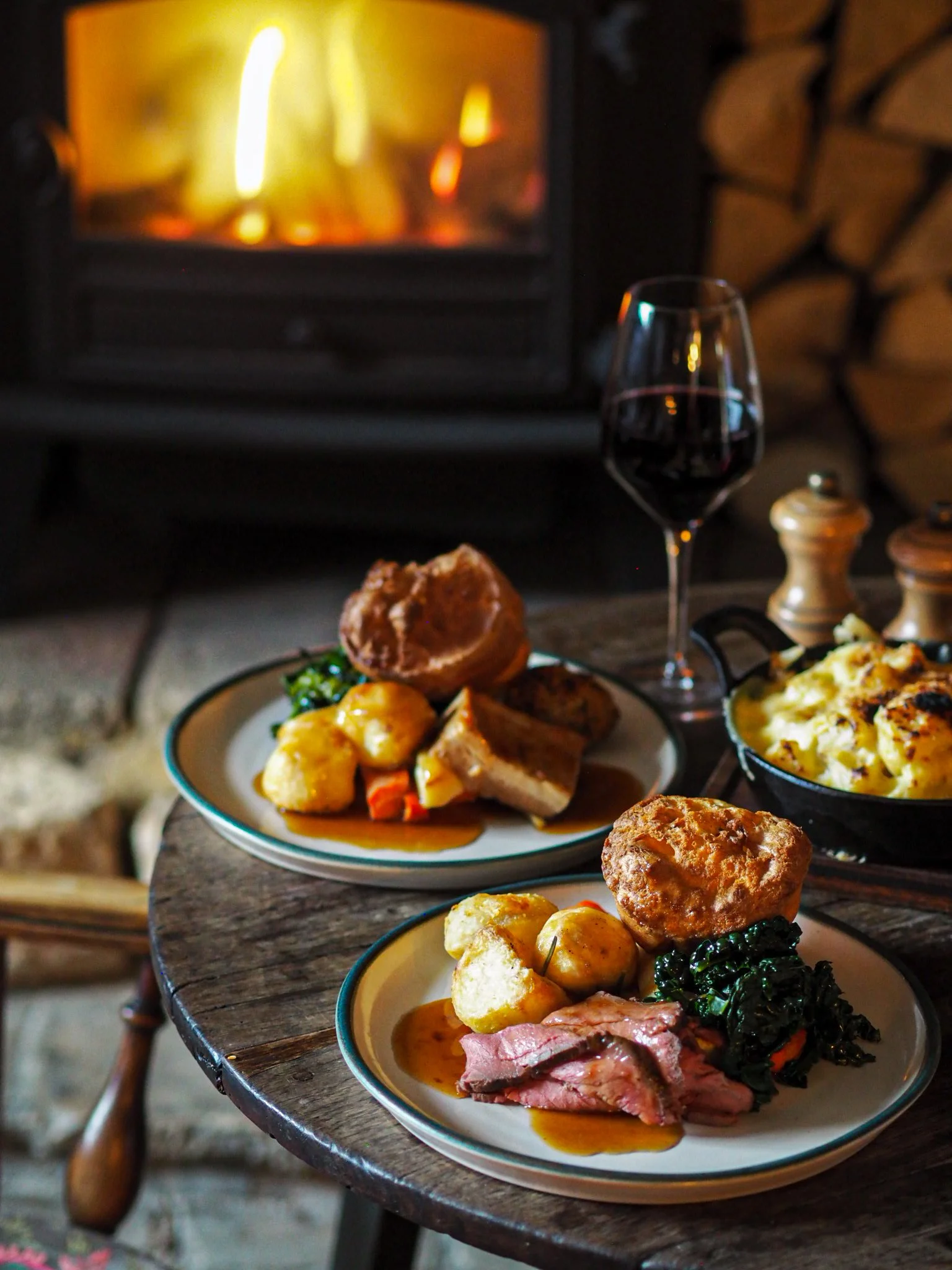Two plates of roast dinner with potatoes, vegetables, gravy, meat, a Yorkshire pudding, and a glass of red wine, set on a rustic wooden table with a fireplace in the background.